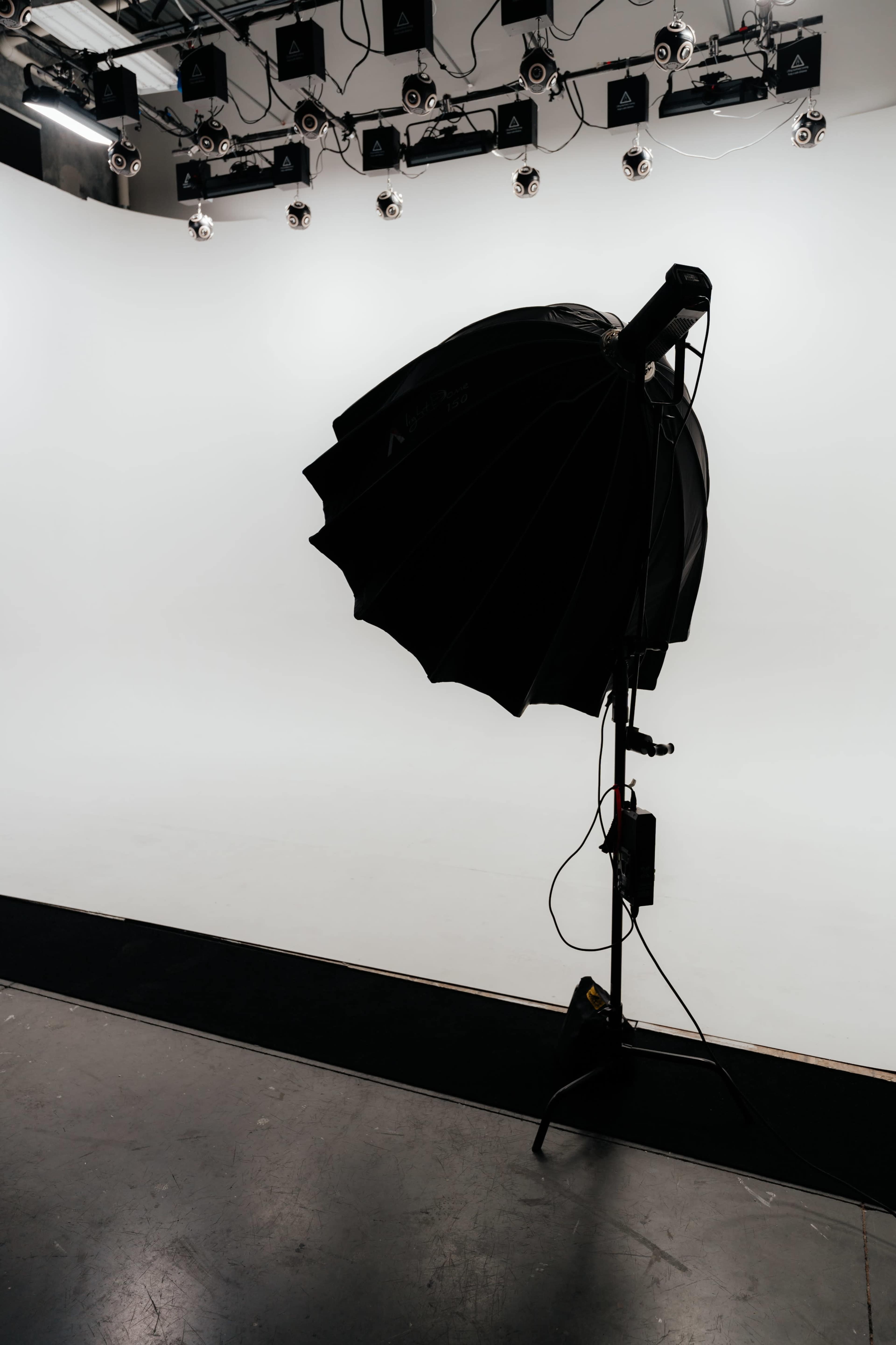 A black softbox light is positioned on a stand in front of a blank white backdrop in a studio setting.