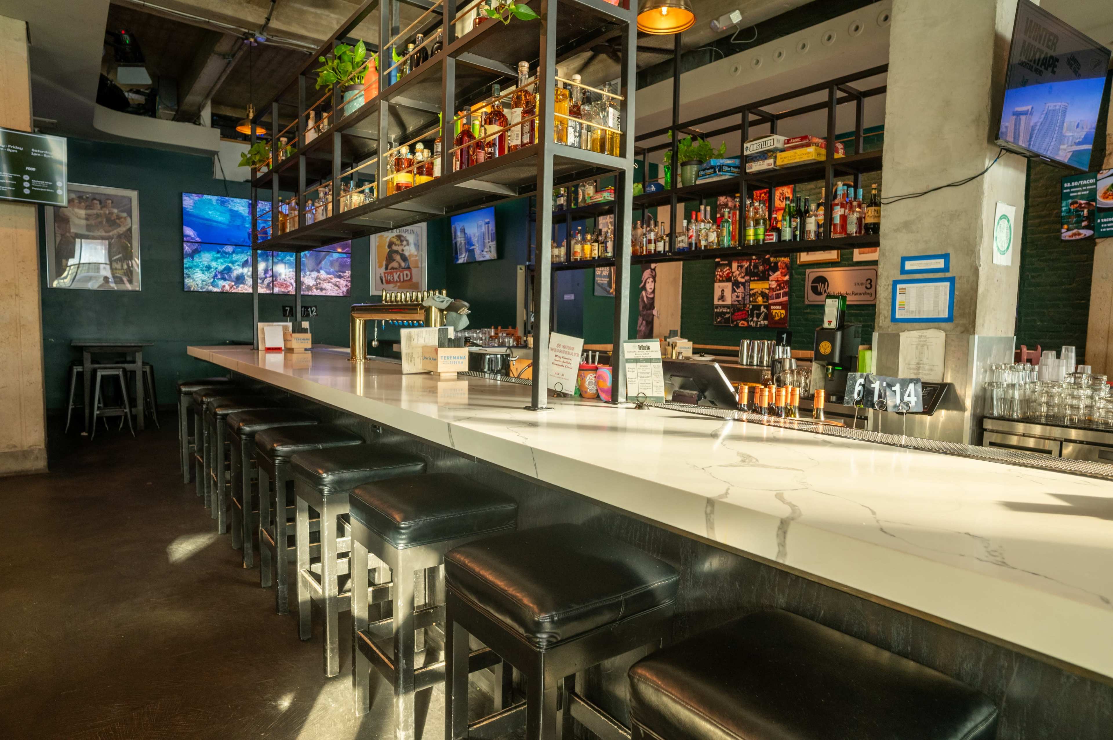 A spacious bar area features a long white countertop with black stools and shelves stocked with various beverages, illuminated by overhead lights.