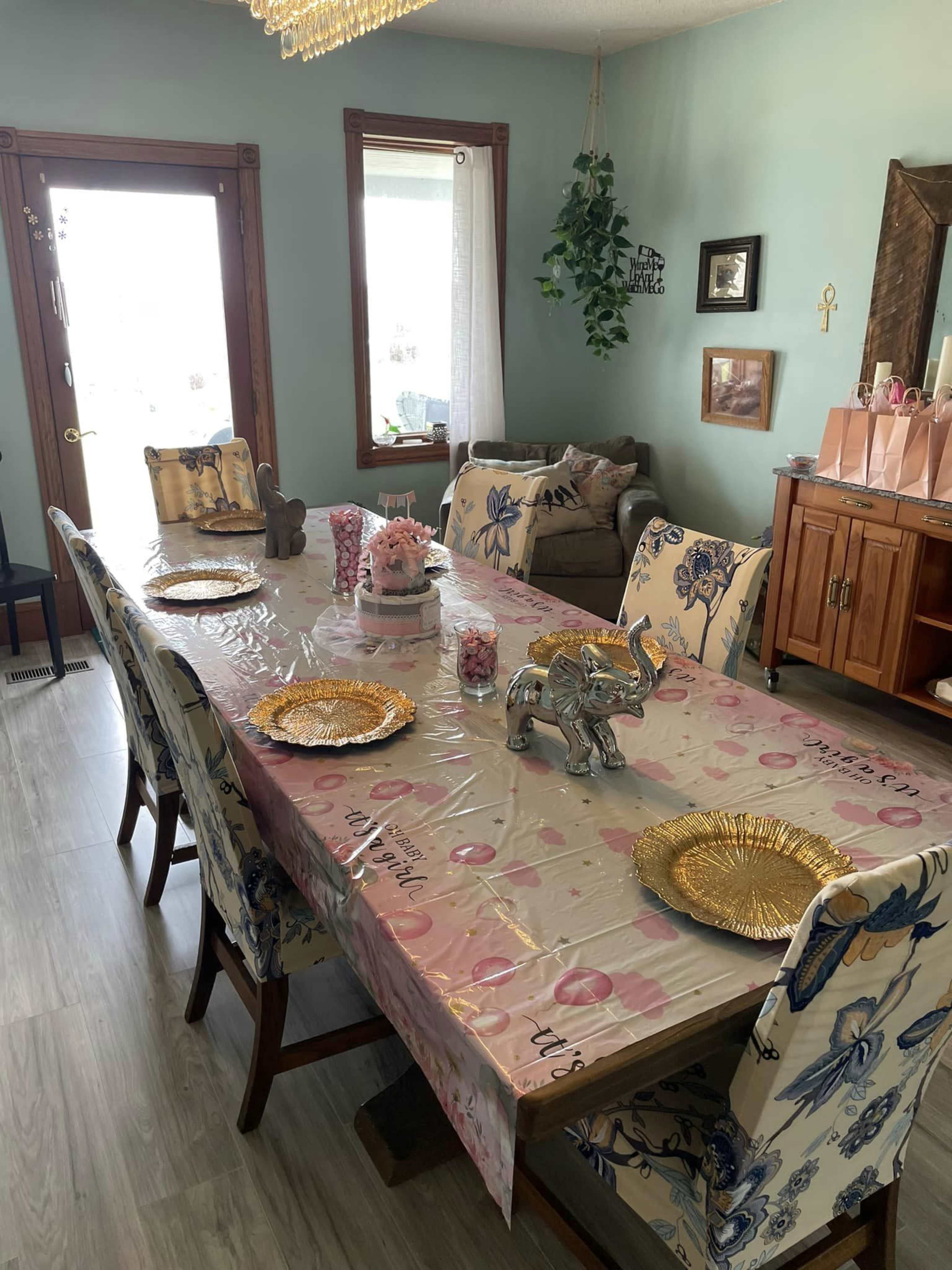 A long dining table is set with gold plates and decorative elephants, surrounded by patterned chairs, in a brightly lit room with a green wall and a large window.