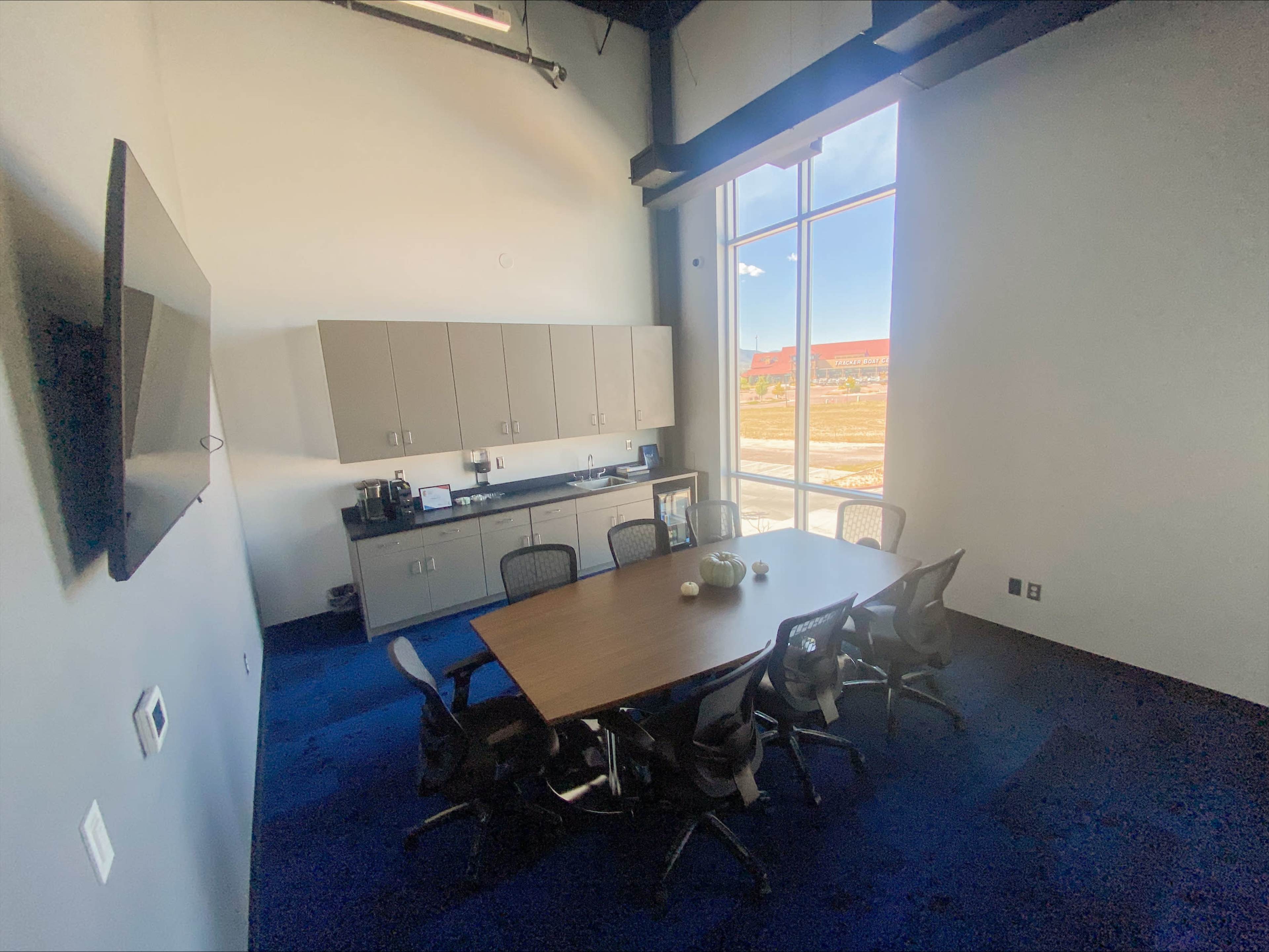 A modern conference room features a large table surrounded by chairs, with a cabinet and kitchen area along one wall and large windows allowing natural light.