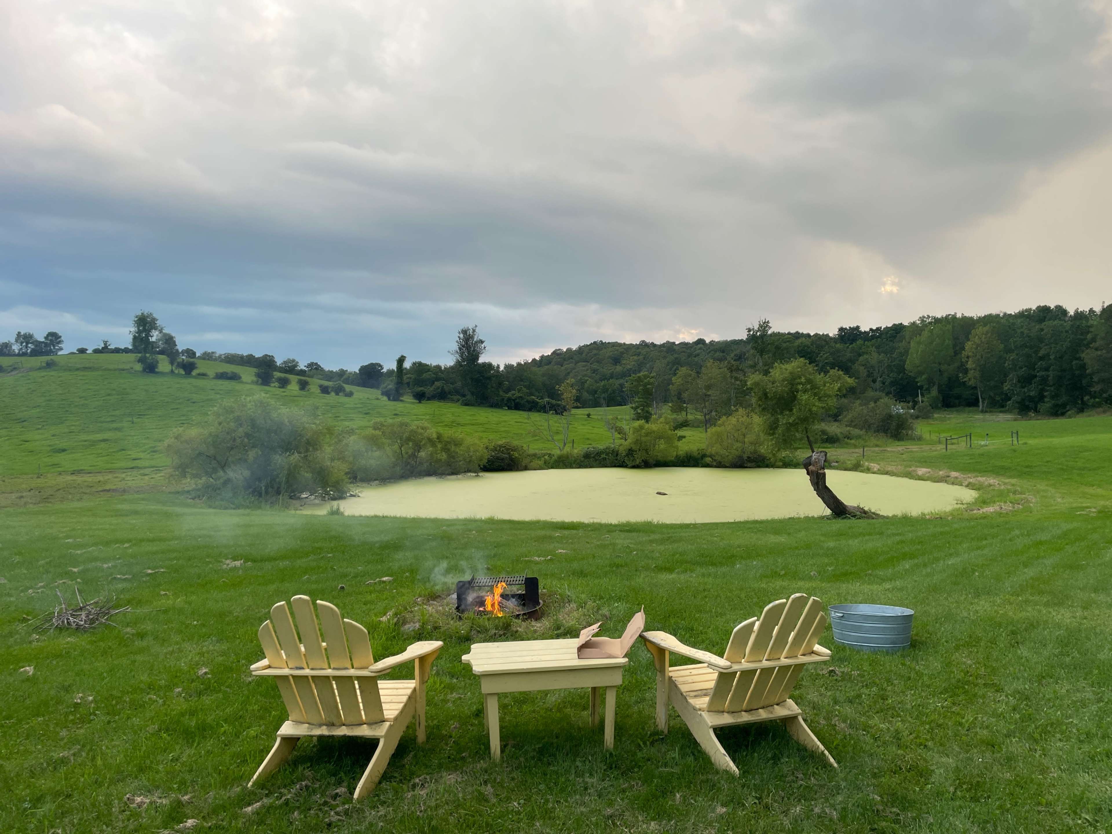 Two wooden chairs and a small table sit beside a pond covered in green algae, with a fire pit burning in front of them on a grassy field.