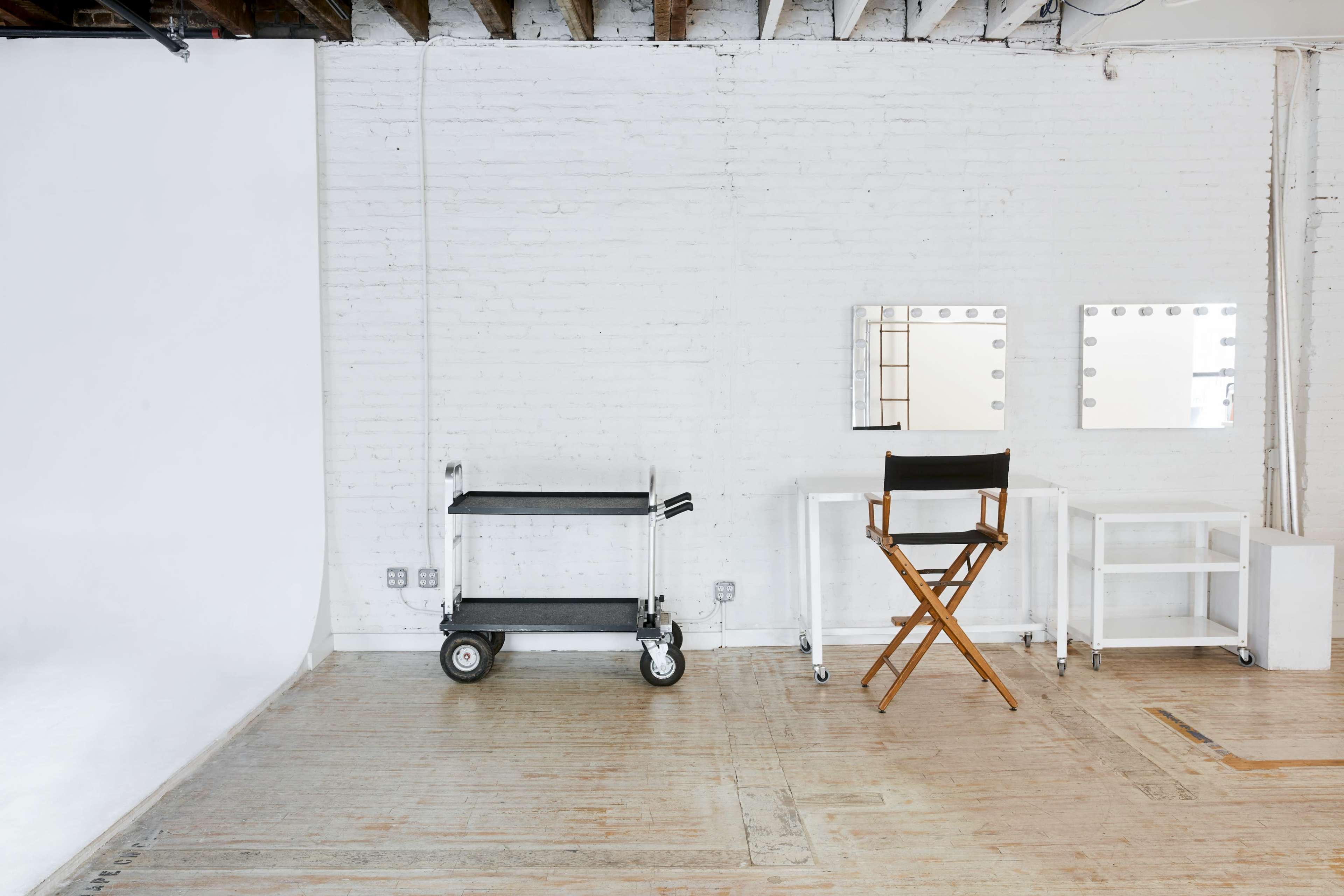 A well-lit studio space features a white backdrop, a cart with wheels, a makeup station with lights, and a director's chair.