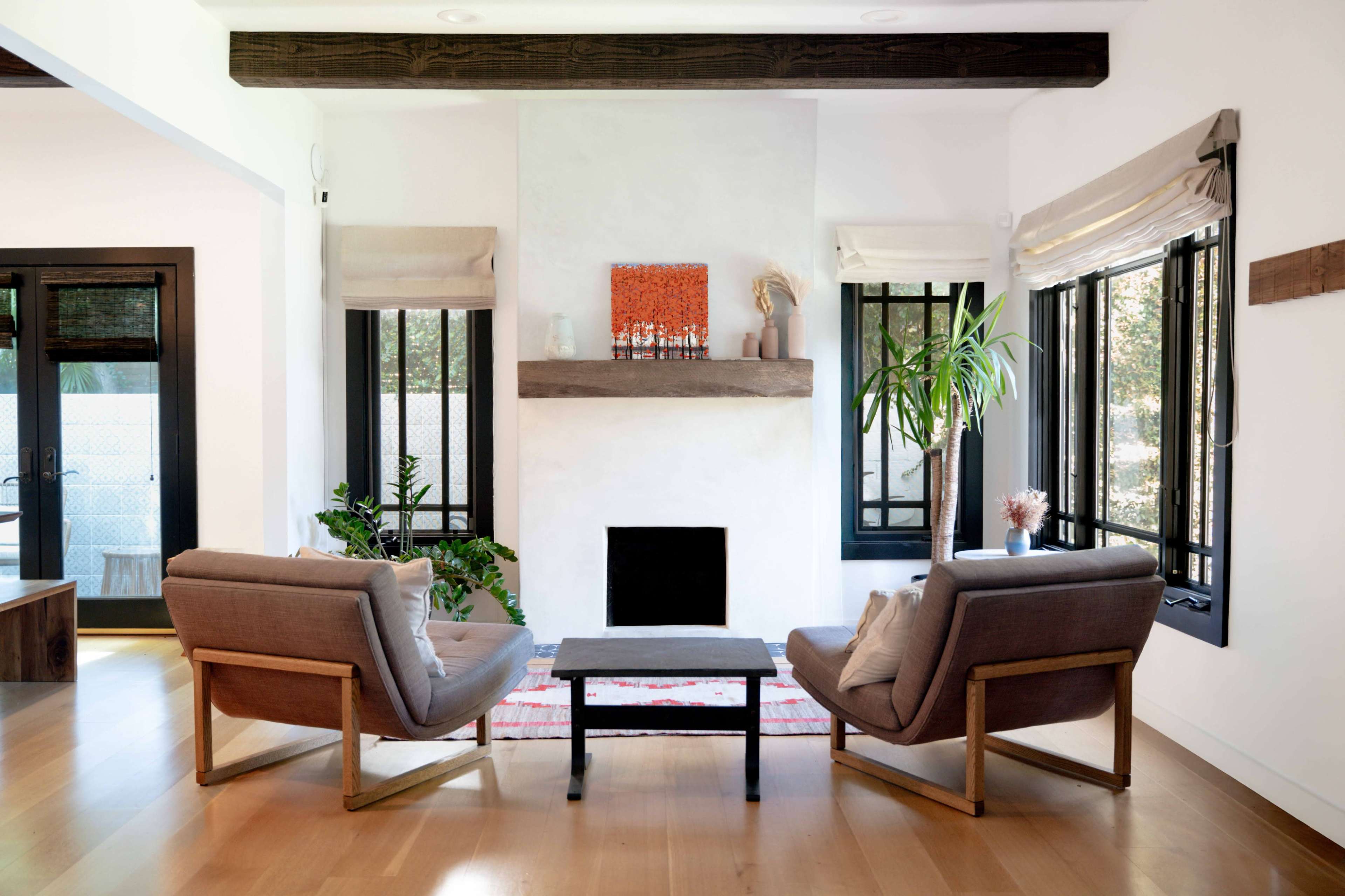 The image depicts a modern living room with two armchairs facing a minimalist fireplace, surrounded by large windows and greenery.