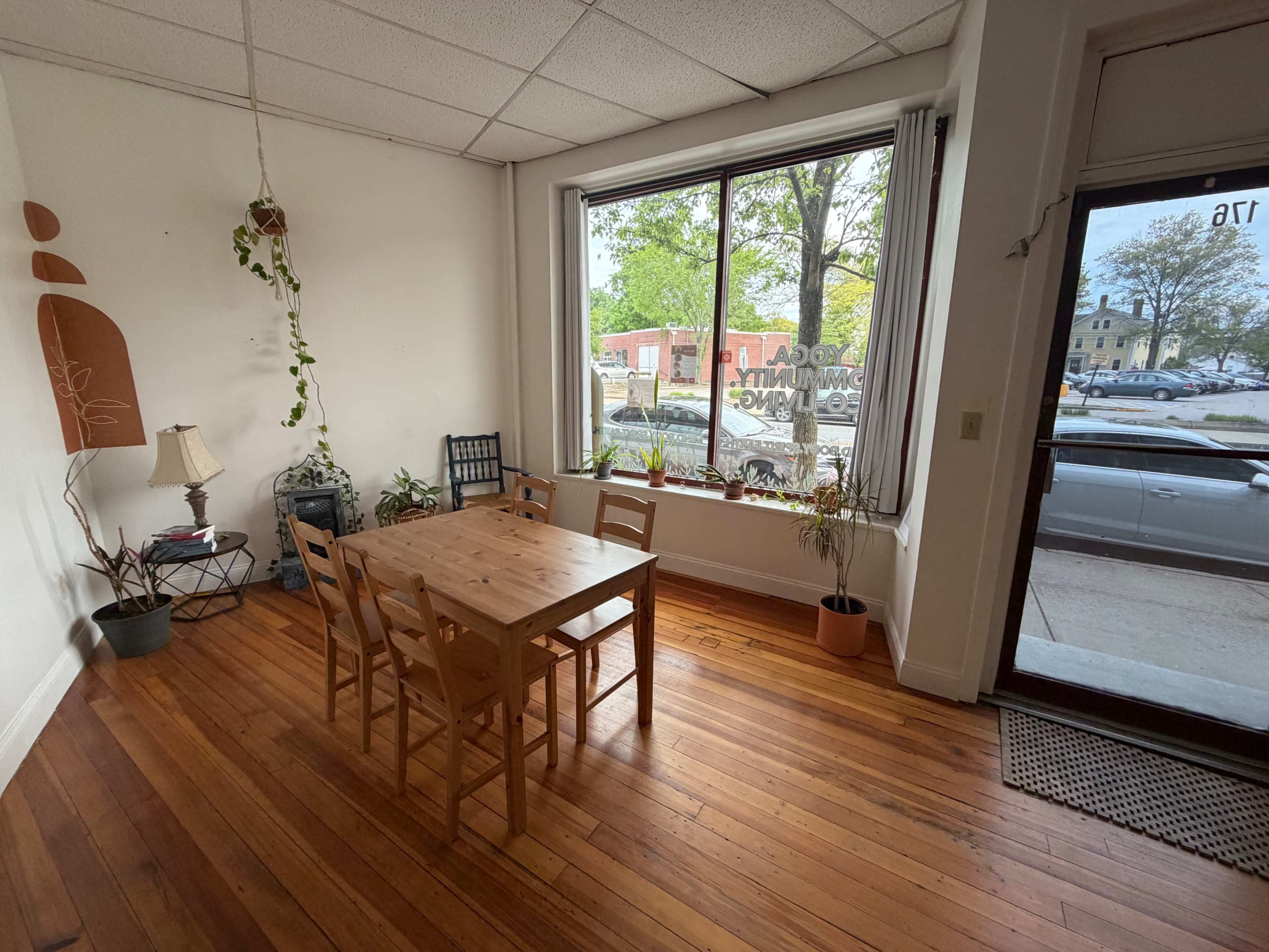 The image shows a small room with a wooden table and four chairs arranged in front of a large window, allowing natural light to enter.