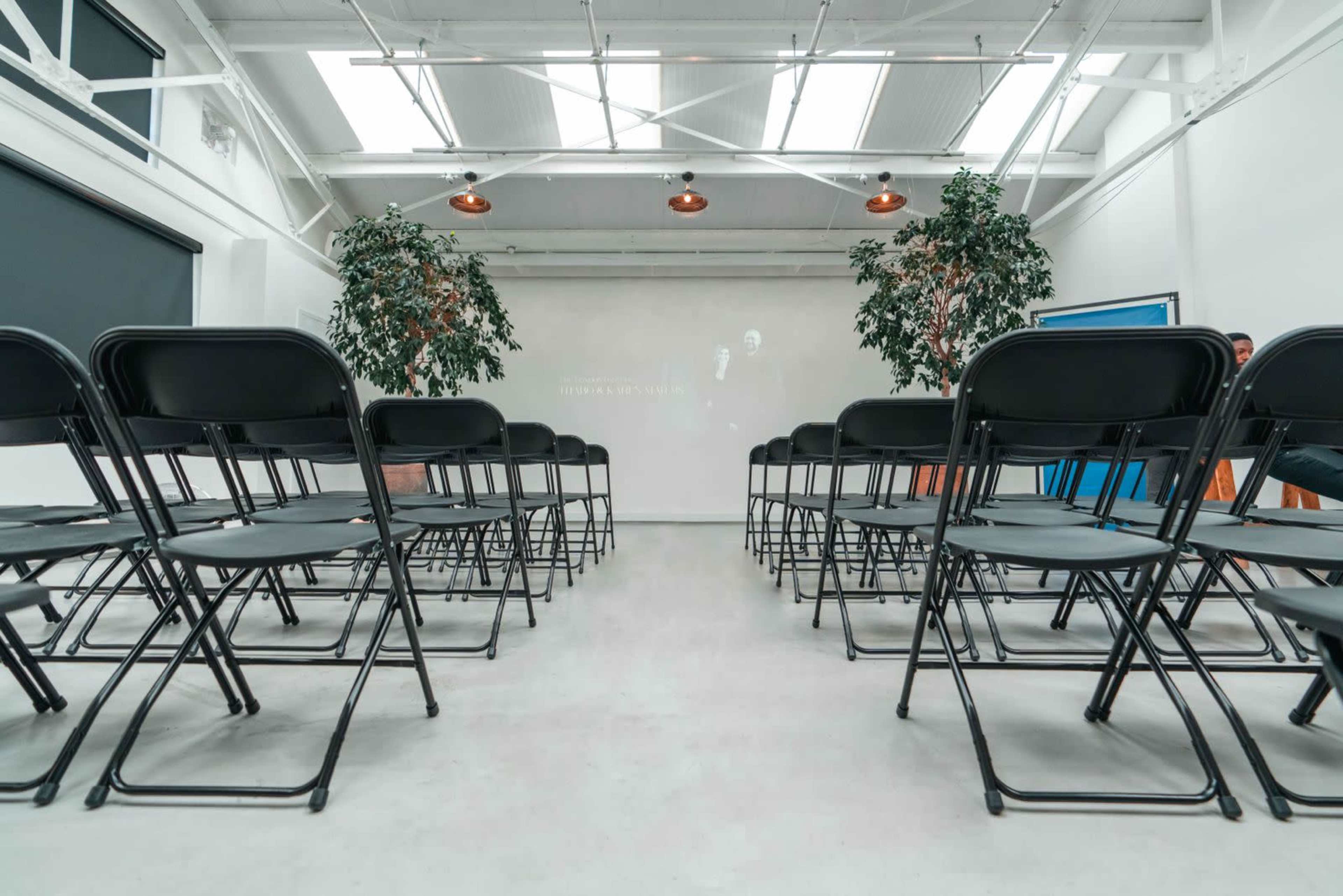 A row of black folding chairs is arranged facing a blank screen in a spacious, well-lit room with potted plants.