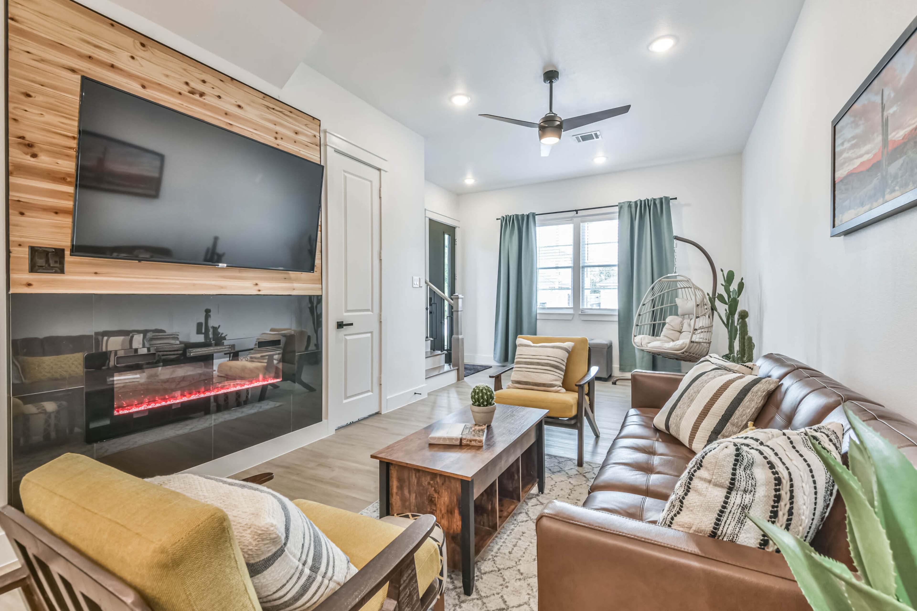 A cozy living room features a wall-mounted TV above an electric fireplace, with a brown leather sofa, two yellow chairs, and large windows covered with light curtains.