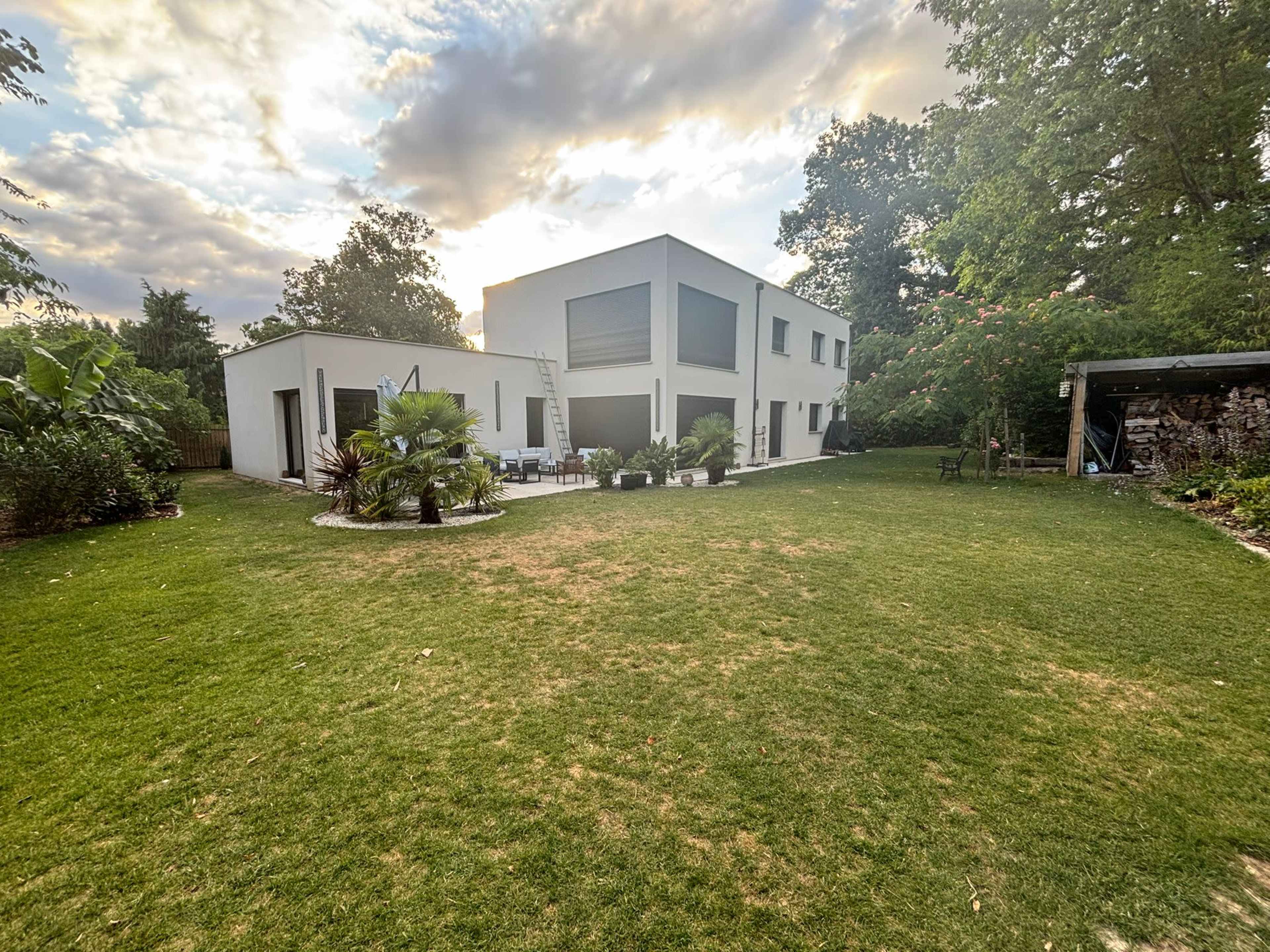 The image shows a modern two-story house set in a spacious green yard with various plants and outdoor furniture.