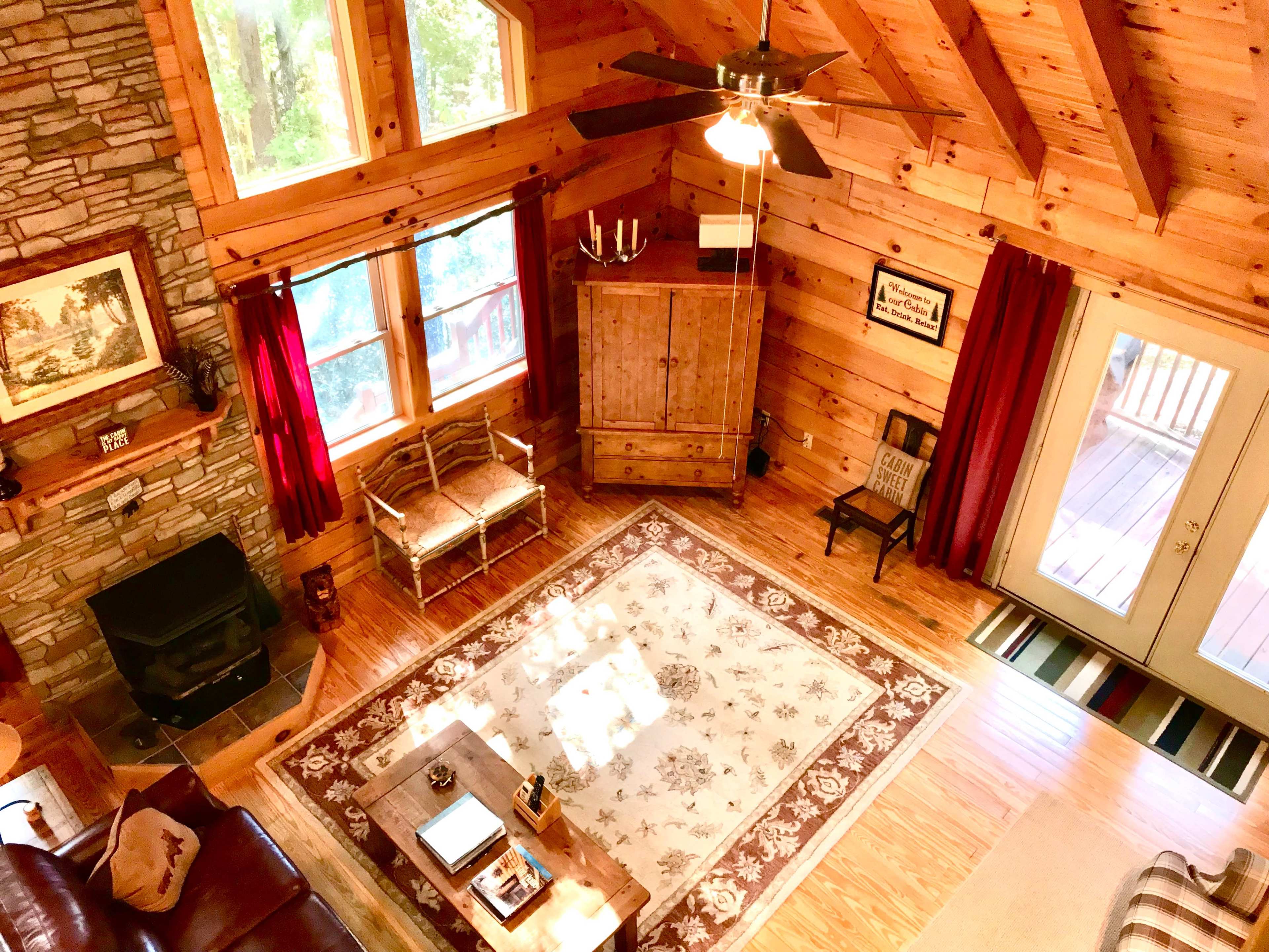 A cozy wooden living room with a stone fireplace, large windows, and a central area rug, furnished with a sofa, chairs, and a coffee table.