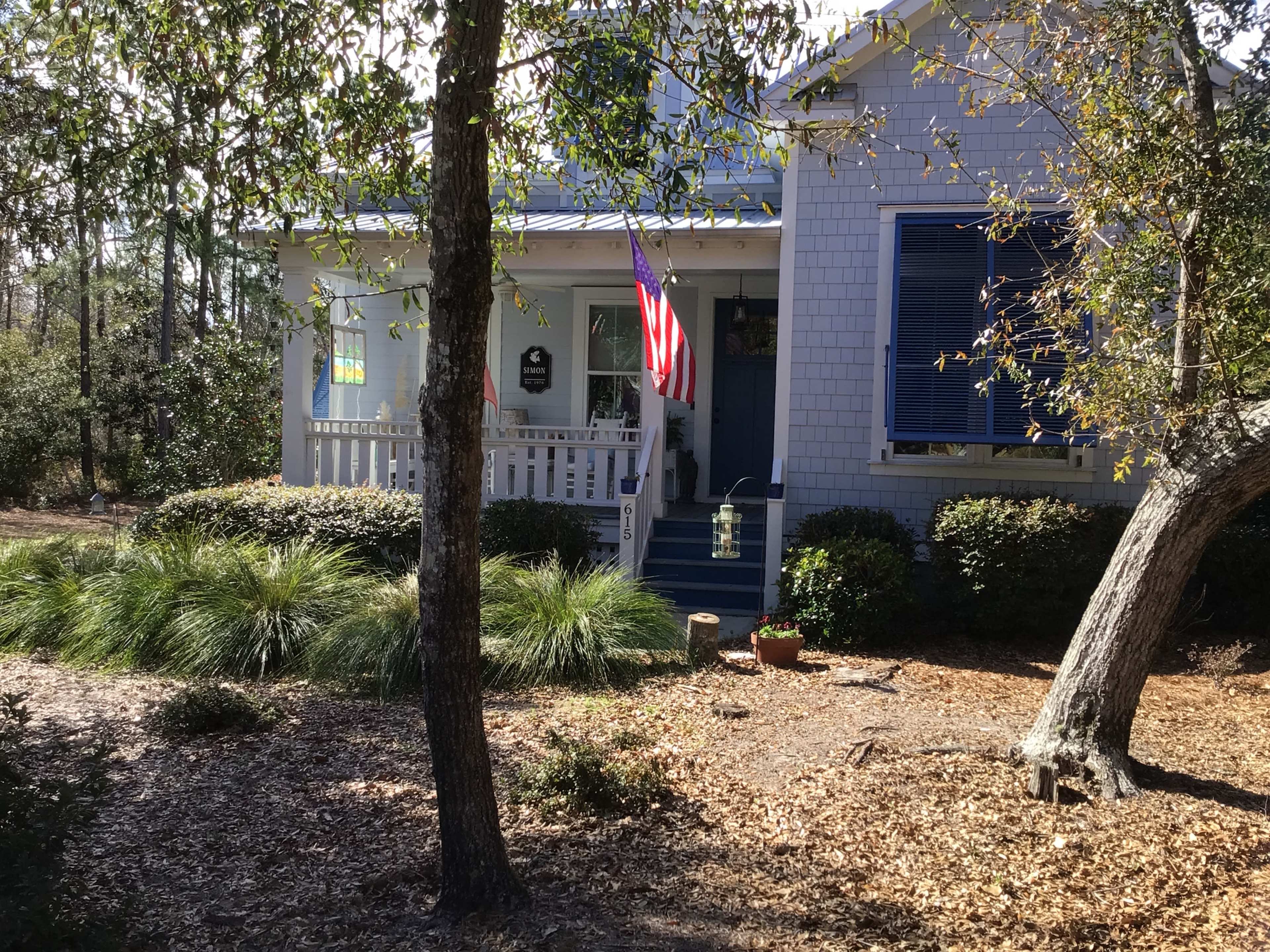 A white house with blue shutters and an American flag displayed on the front porch is surrounded by greenery and mulch.