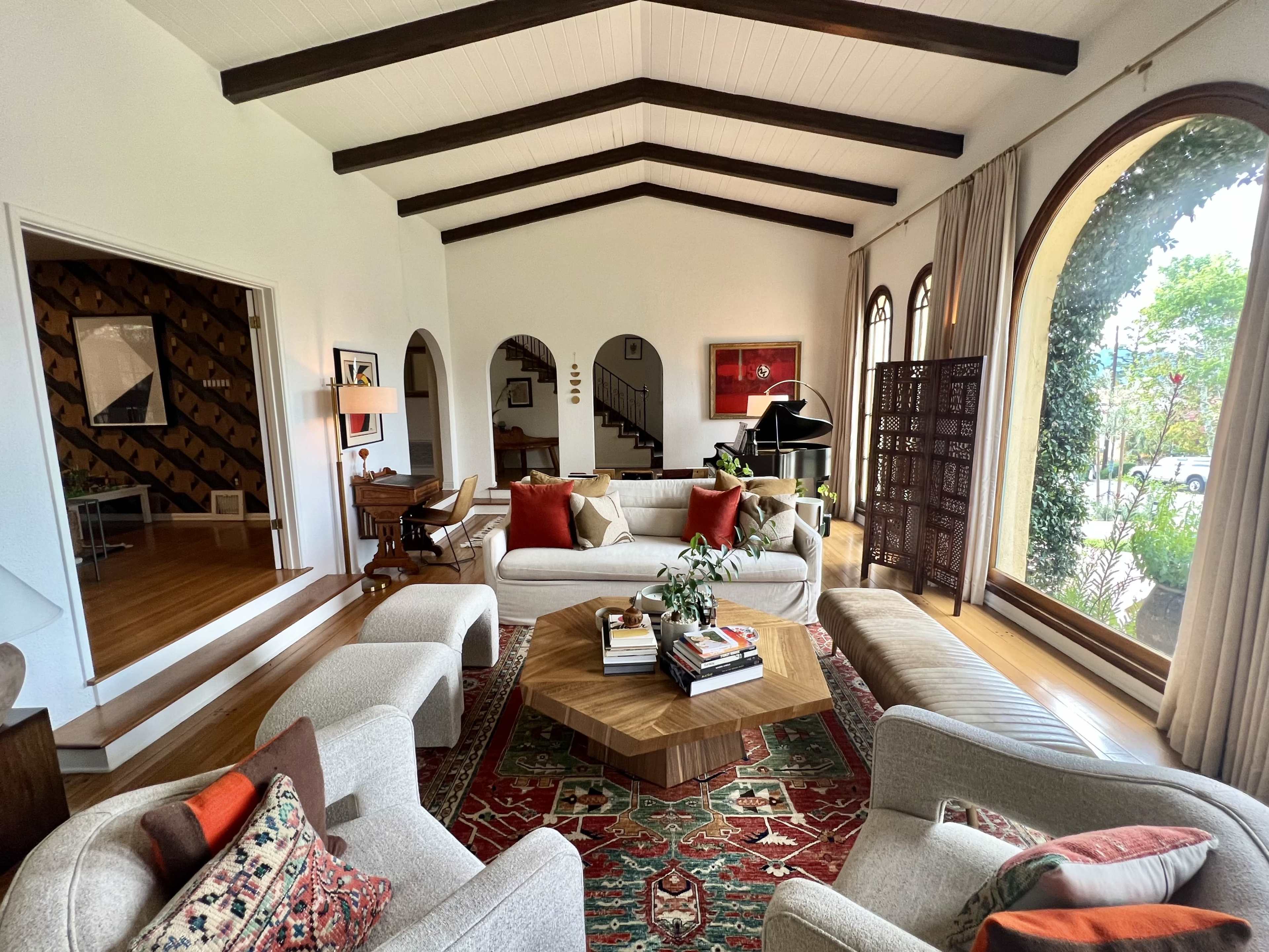 A spacious living room with high wooden-beamed ceilings, featuring two light-colored sofas, an octagonal coffee table, and large windows that overlook a garden.