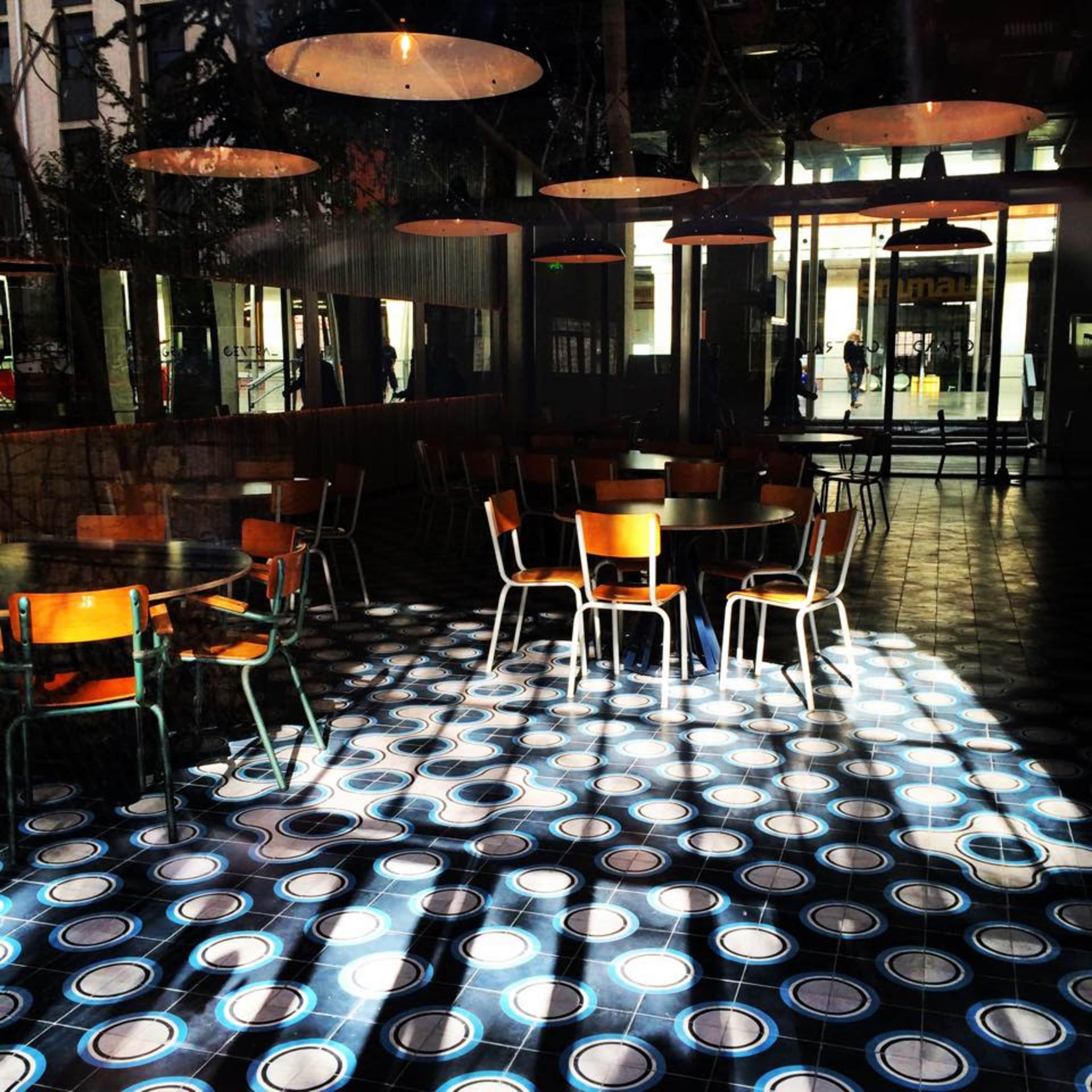 An empty café with wooden chairs and tables, reflecting sunlight on a patterned tiled floor.