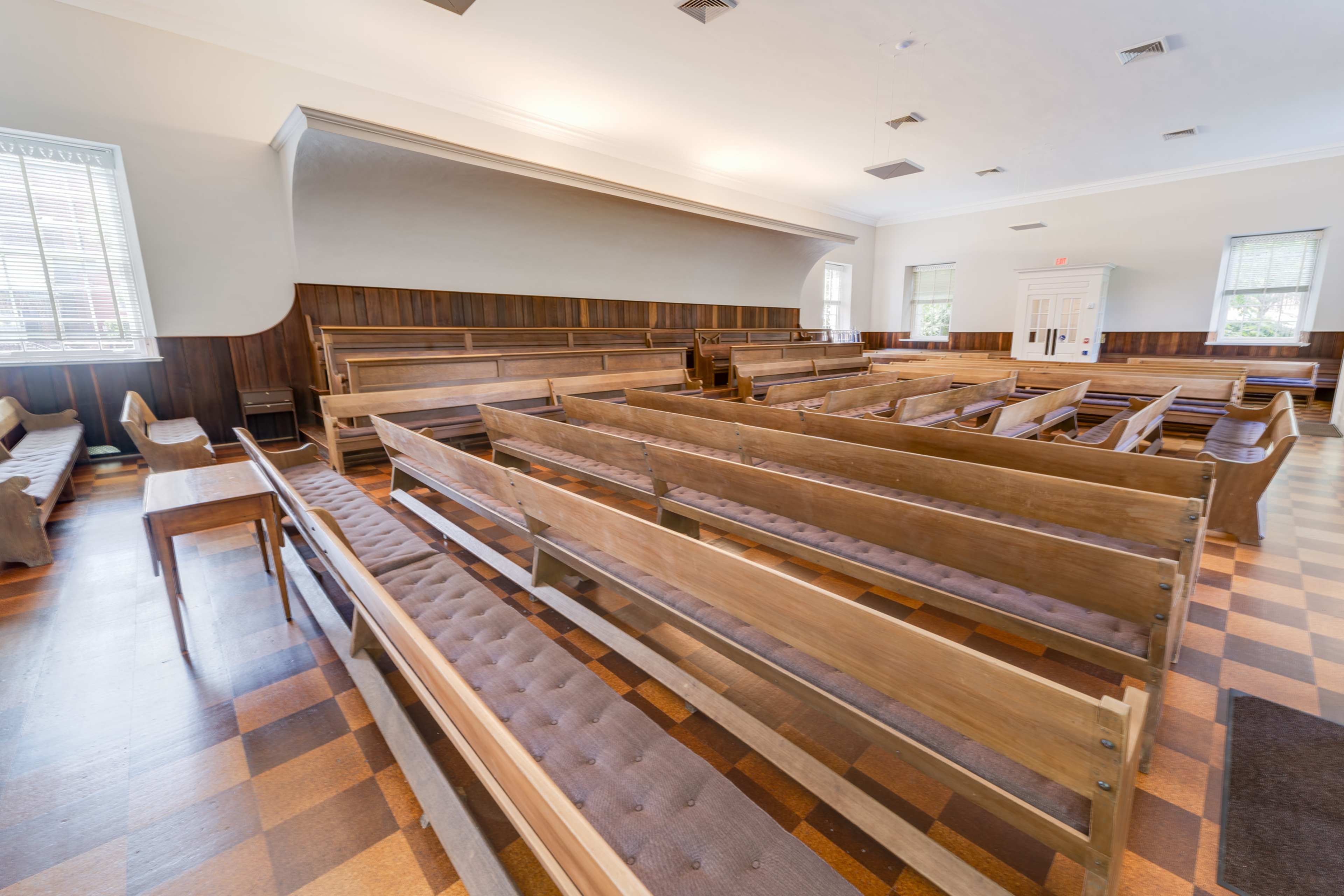 A spacious interior of a room with wooden benches arranged in rows on a checkered floor.