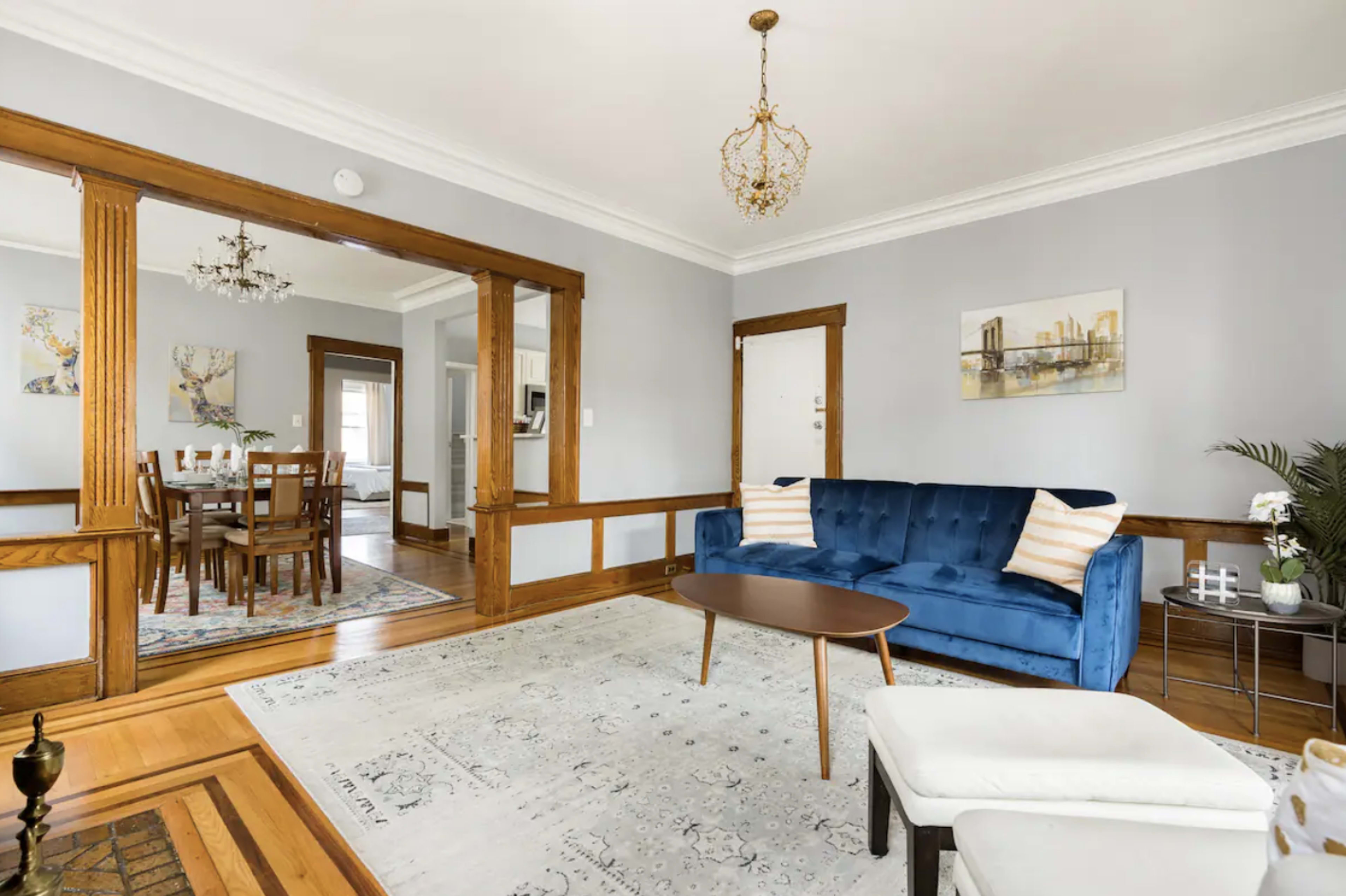 A living room with a blue velvet sofa, a round wooden coffee table, and a light-colored area rug, featuring wood trim and chandeliers.