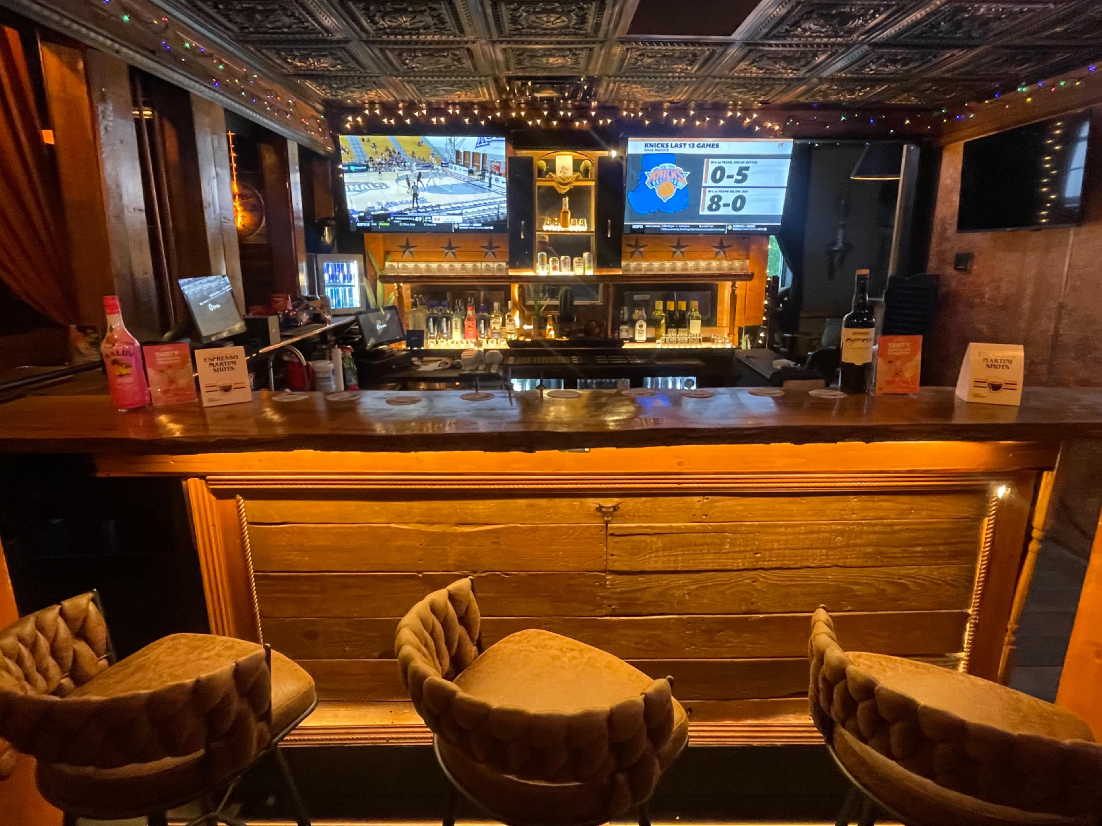 The image shows a bar with a wooden counter, three upholstered stools, and two televisions displaying sports scores.