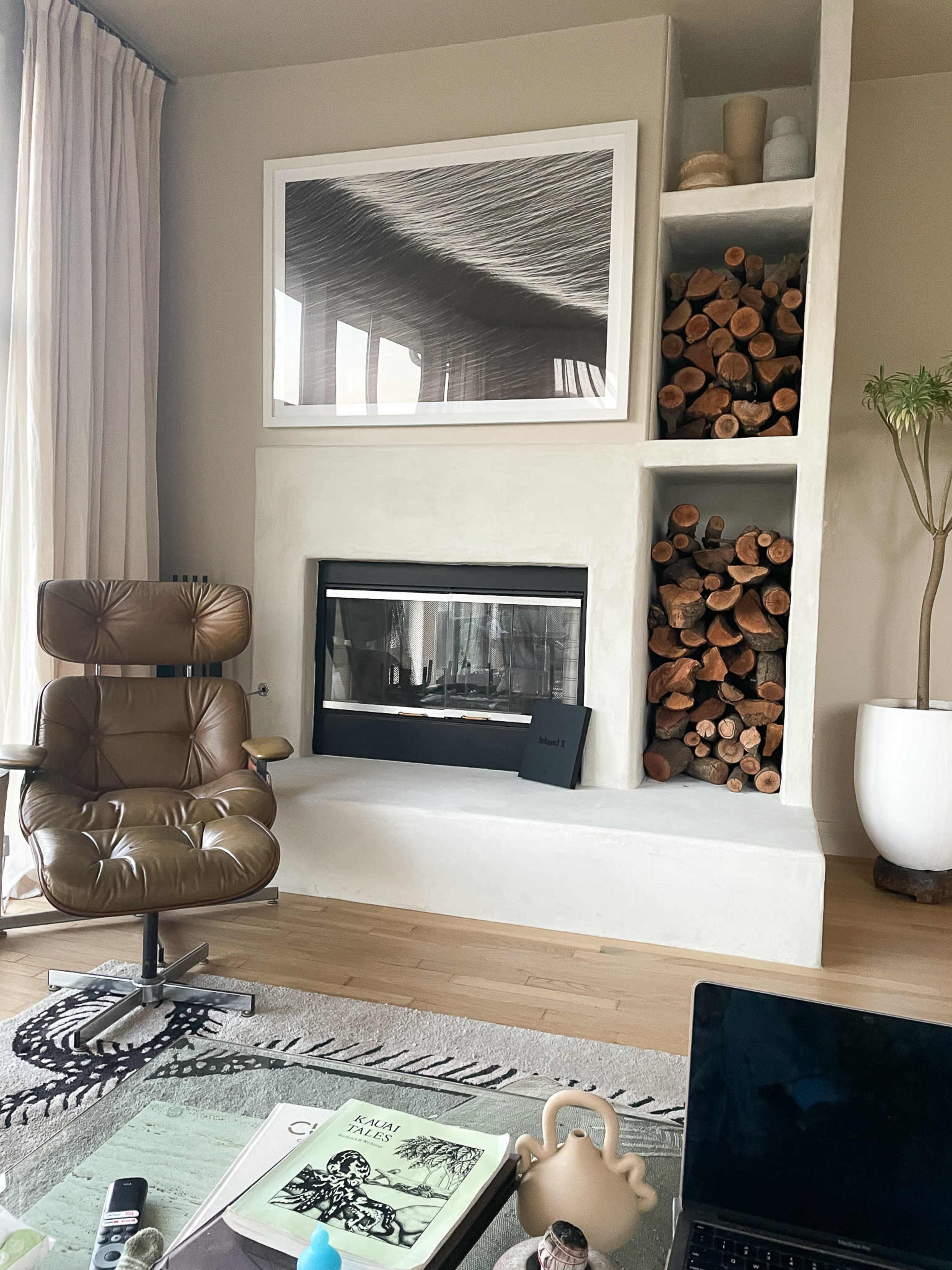 A modern living room with a fireplace built into a wall lined with stacked firewood and a brown armchair nearby.