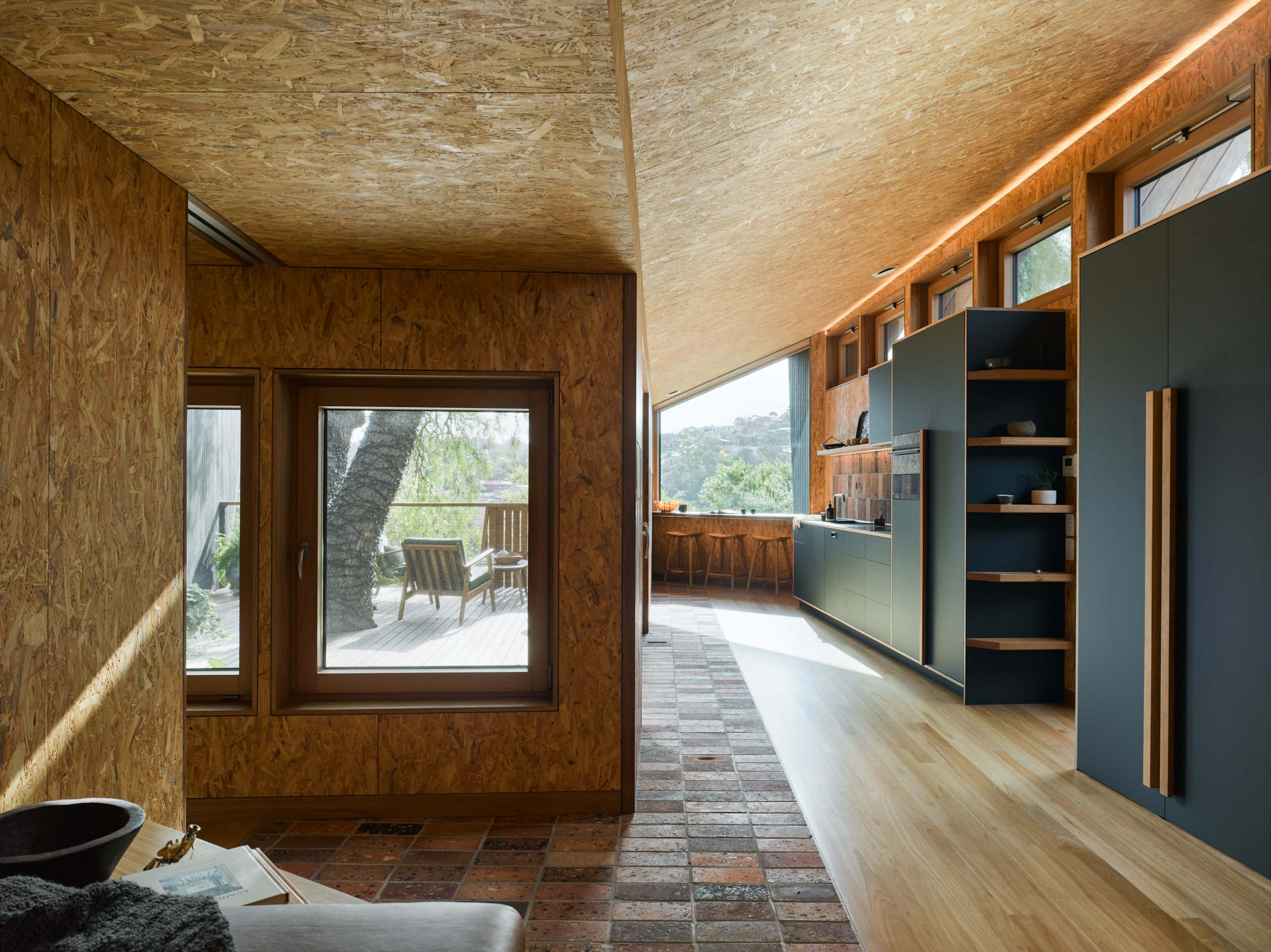 The image shows an interior space with wooden walls and large windows, leading to a modern kitchen area that features sleek cabinetry and a view of an outdoor deck.