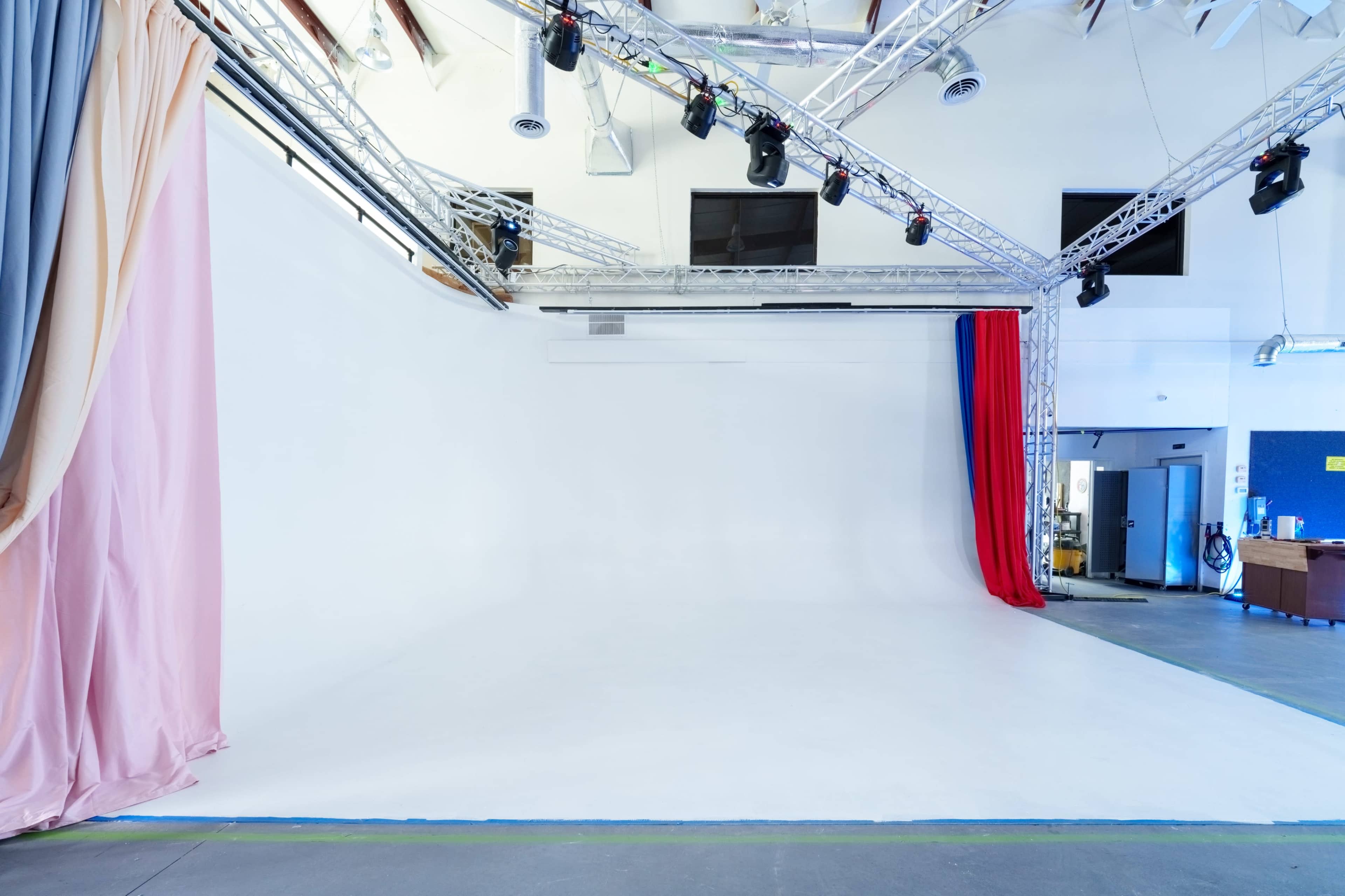 The image shows a large, empty studio space with a white backdrop, soft lighting, and draped fabric in pastel and red colors.