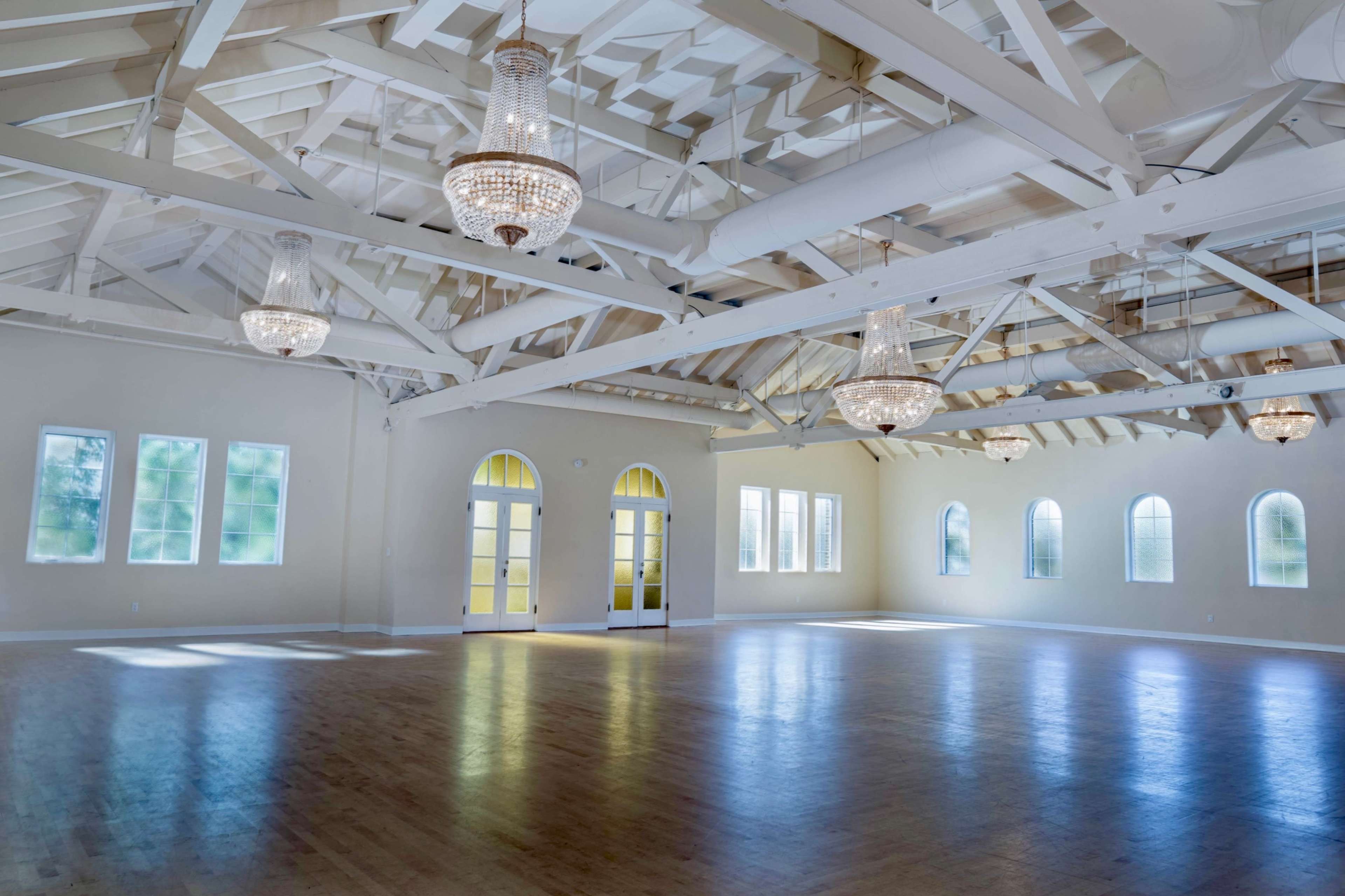 The image shows a spacious, well-lit interior room with a high ceiling, featuring multiple large windows and ornate chandeliers.