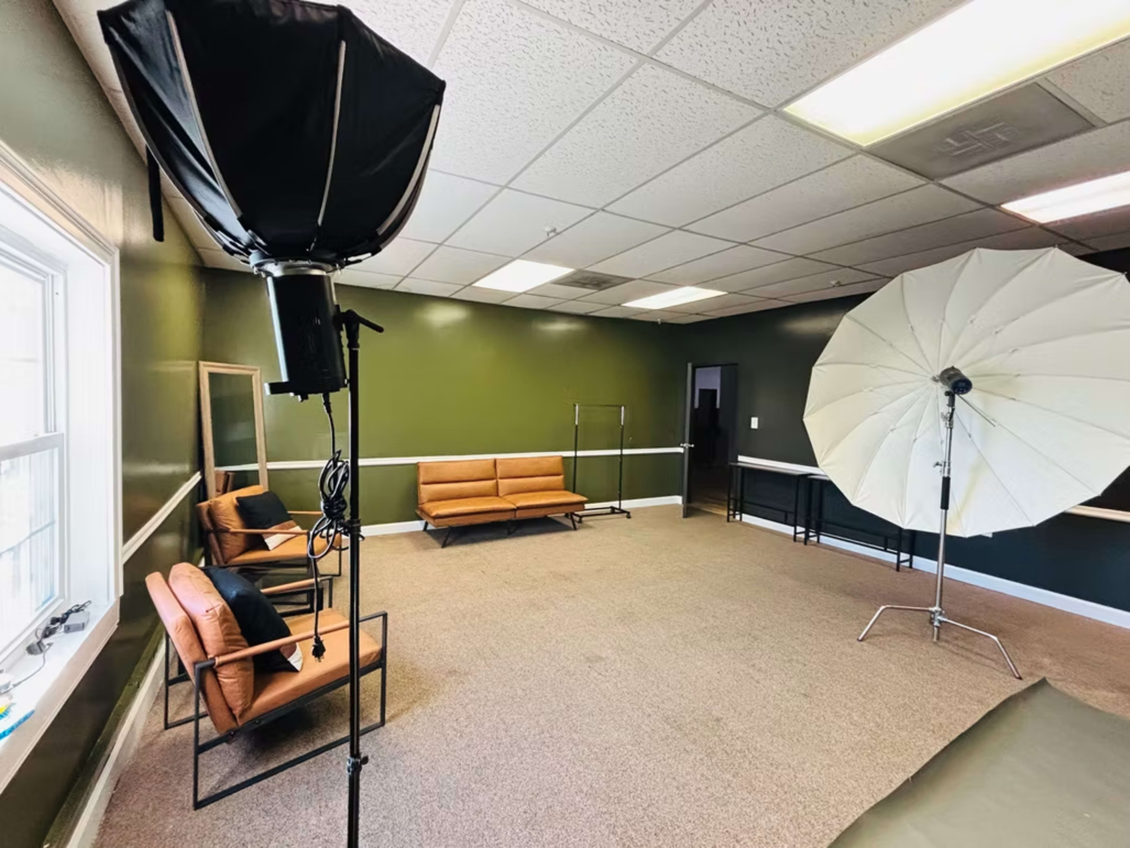 The image shows an empty room with a dark green wall, featuring two brown sofas, a large umbrella softbox, and a standing mirror.