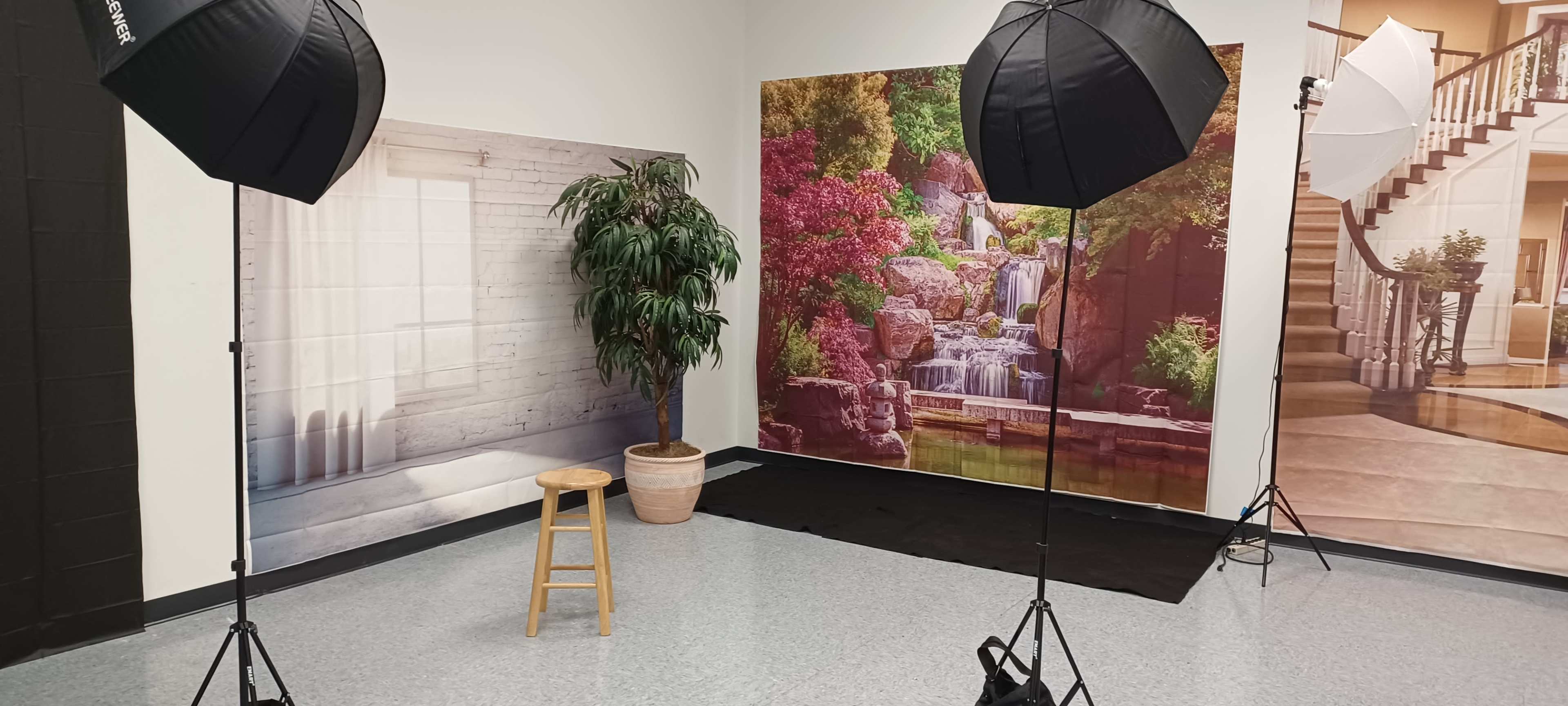 A photography studio setup with two softbox lights, a wooden stool, and colorful backdrop images depicting a waterfall scene and an interior view.