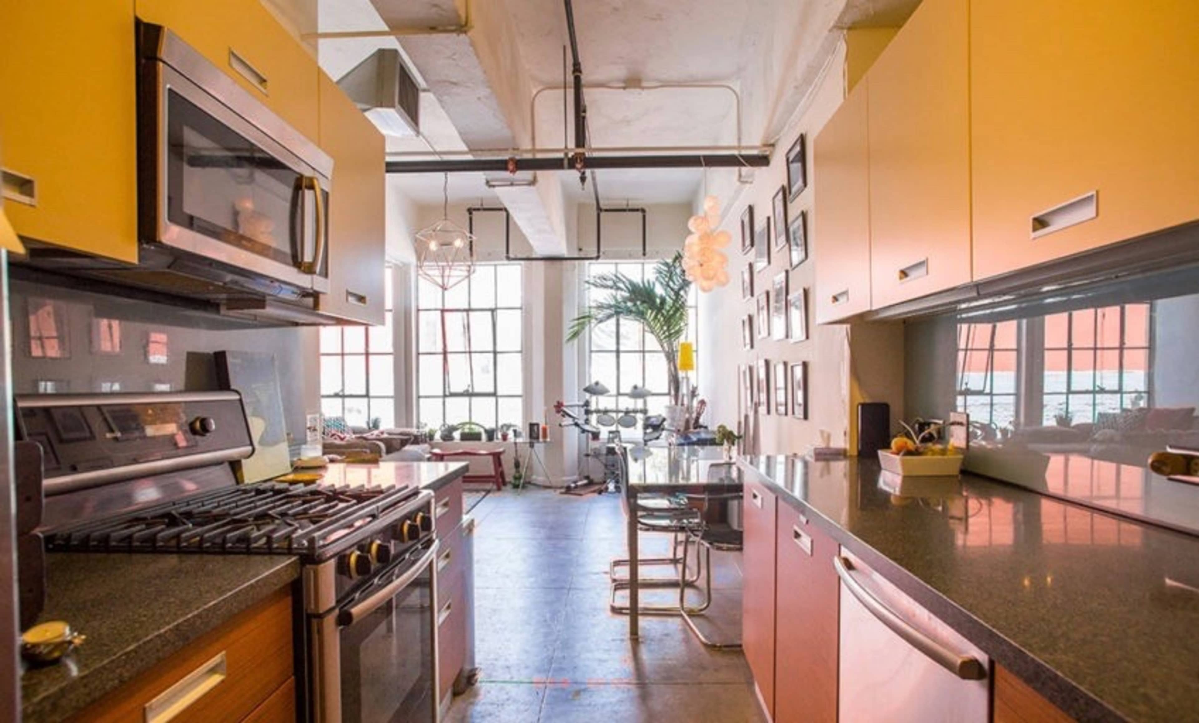 The image shows a modern kitchen with stainless steel appliances, a sleek countertop, and large windows providing natural light to a connected dining area.