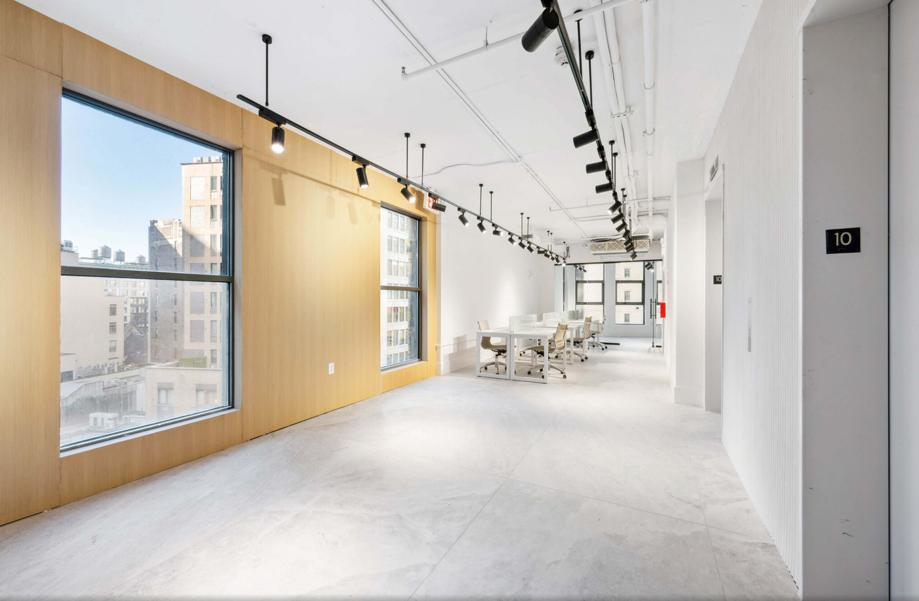 The image shows a bright, modern office space with large windows, light-colored walls, and a meeting area with a long table.