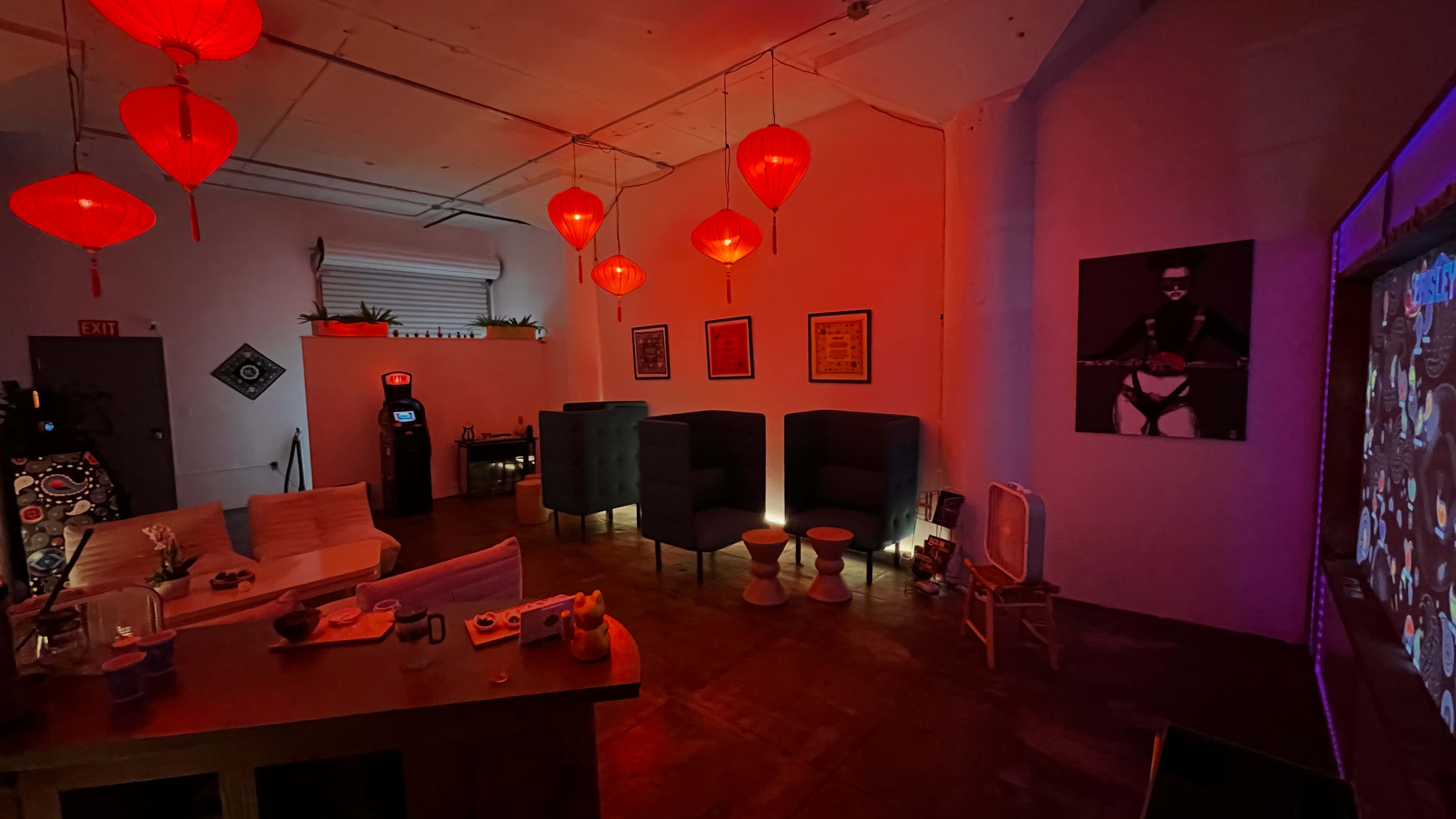 The image shows a dimly lit lounge area with red lanterns hanging from the ceiling, seating arranged around a central bar, and a wall featuring a large artwork.