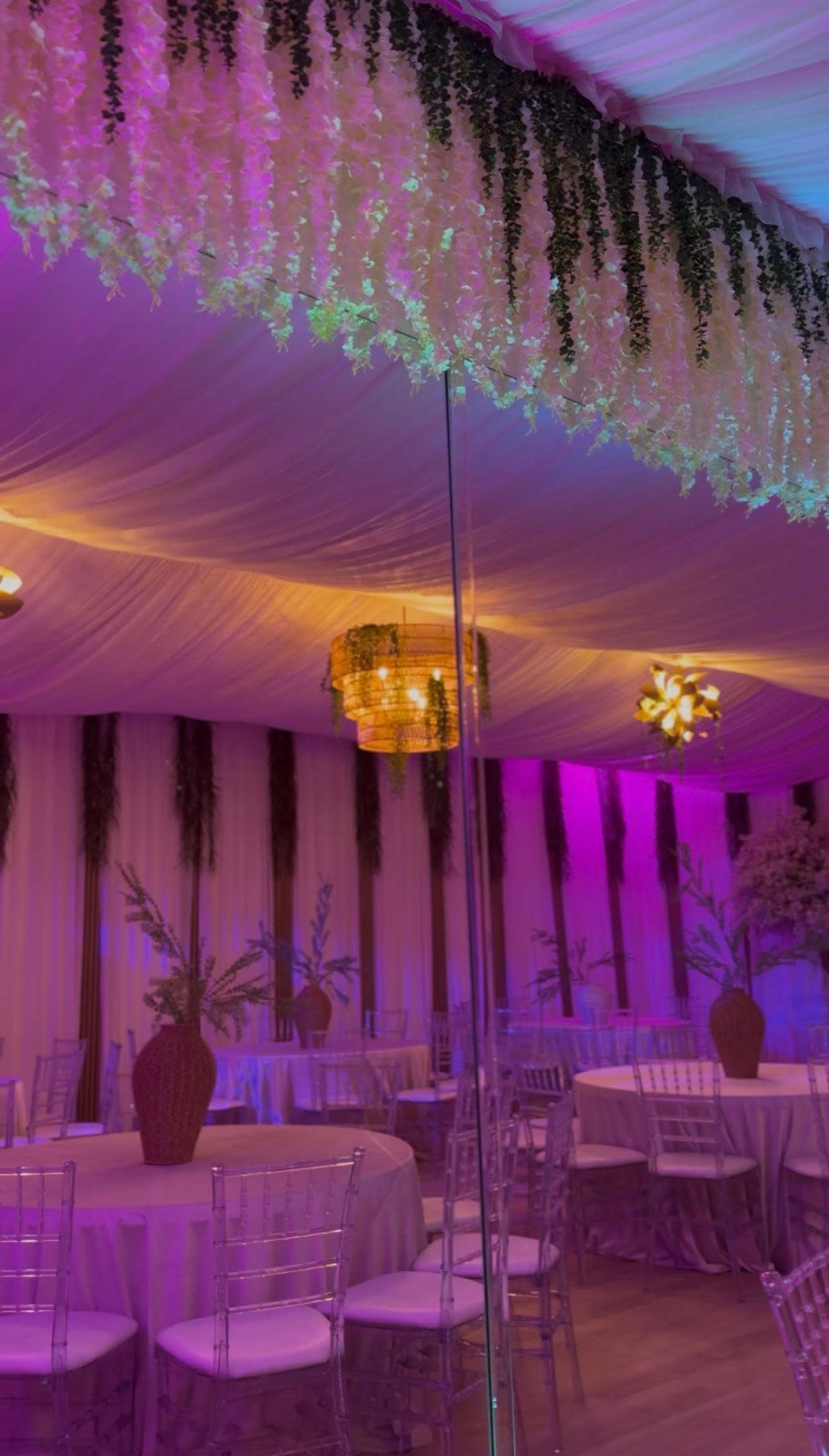 A decorated banquet hall features round tables with white covers, elegant chairs, floral arrangements, and vibrant purple and warm lighting.