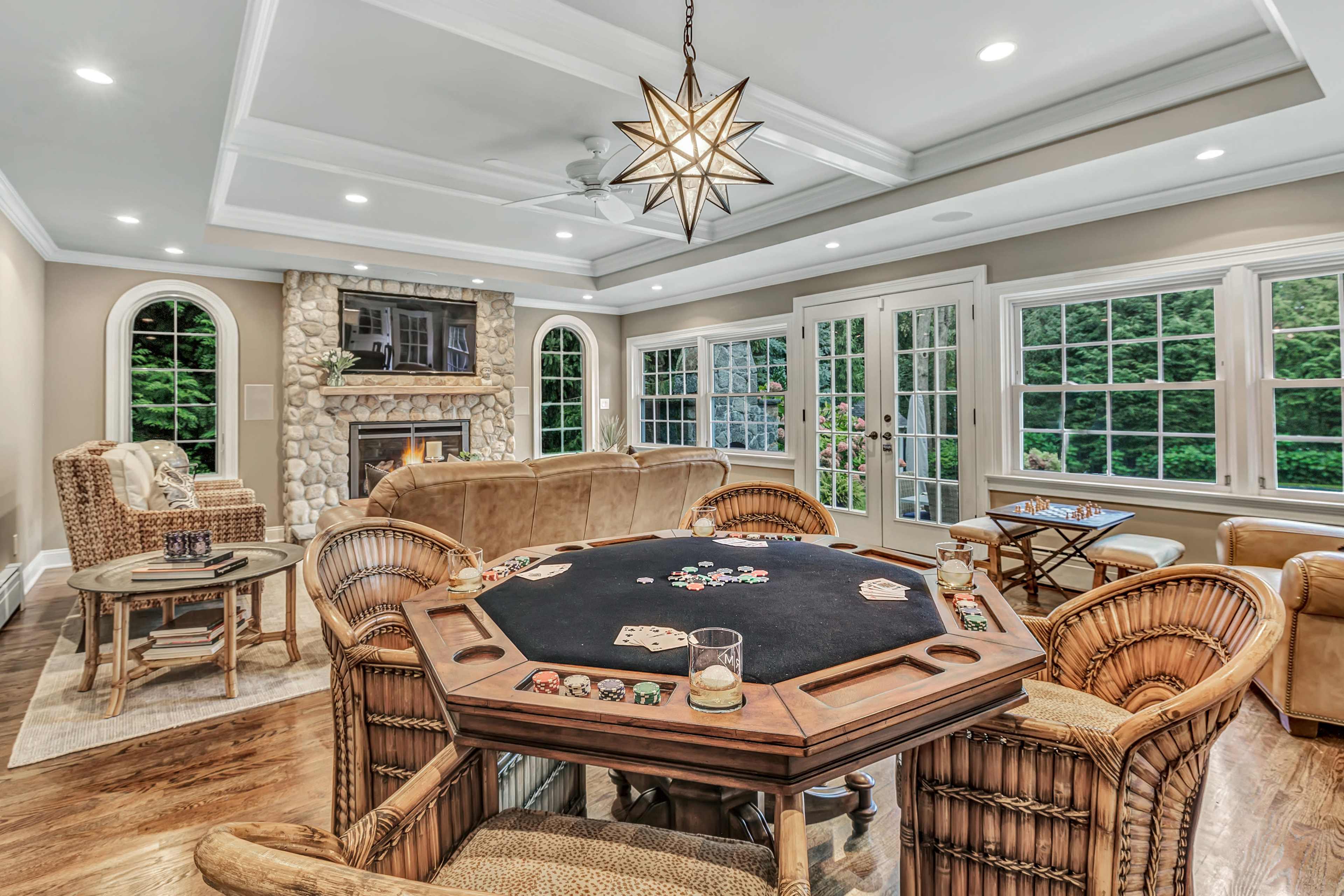 A game room features a octagonal poker table surrounded by wicker chairs, with a fireplace and large windows allowing natural light.
