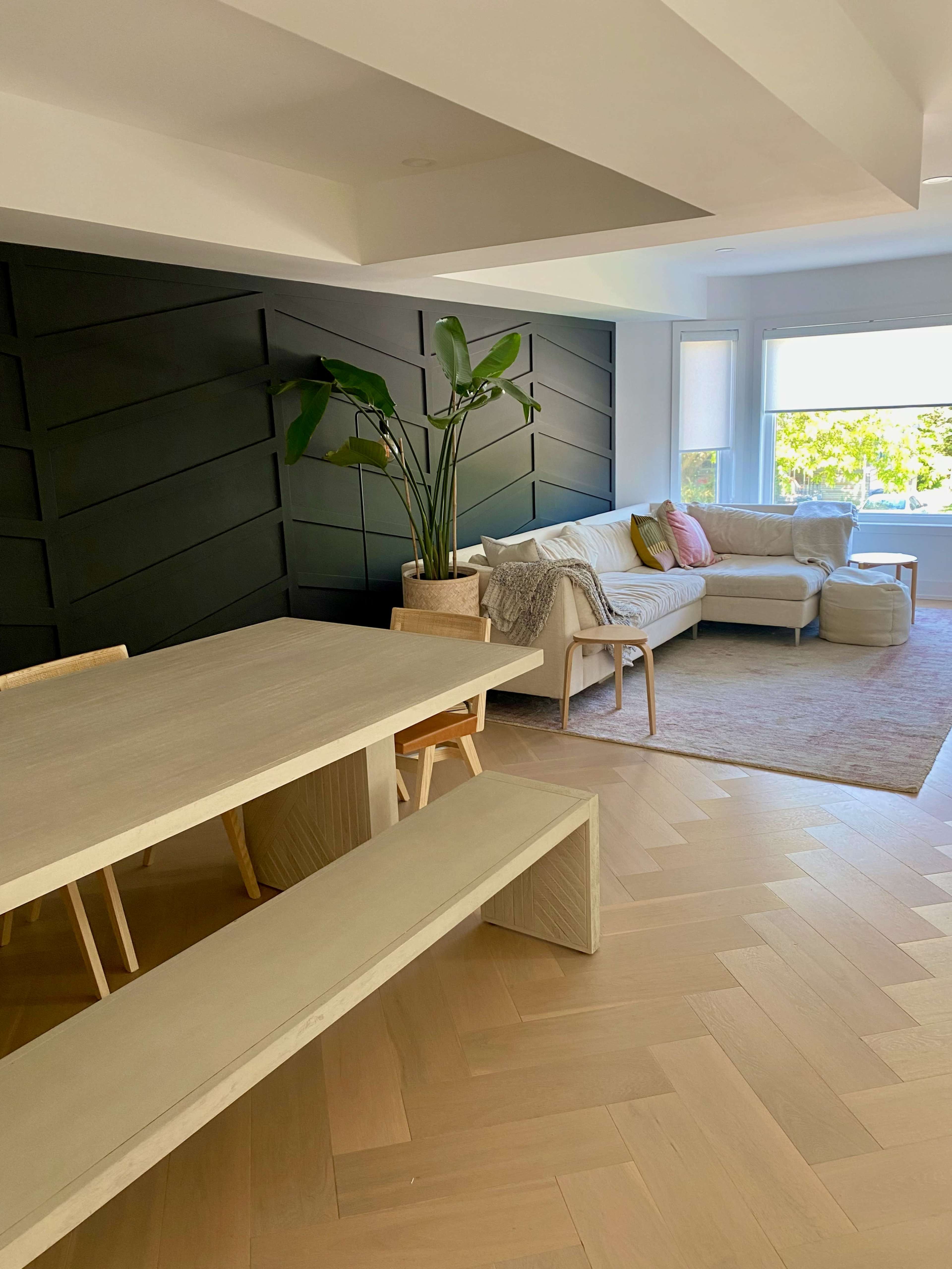 The image shows a modern living and dining area featuring a light wooden dining table with matching benches, a light-colored sofa, and a large window with natural light illuminating the space.