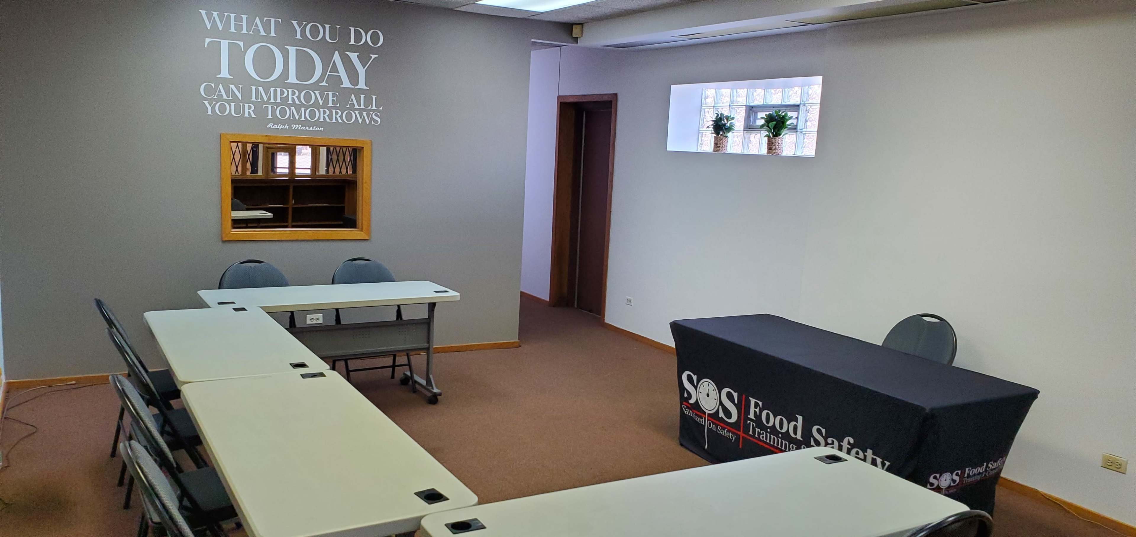 A training room with several tables arranged in a U-shape, a table with a logo for "SAS Food Safety," and a motivational quote on the wall.