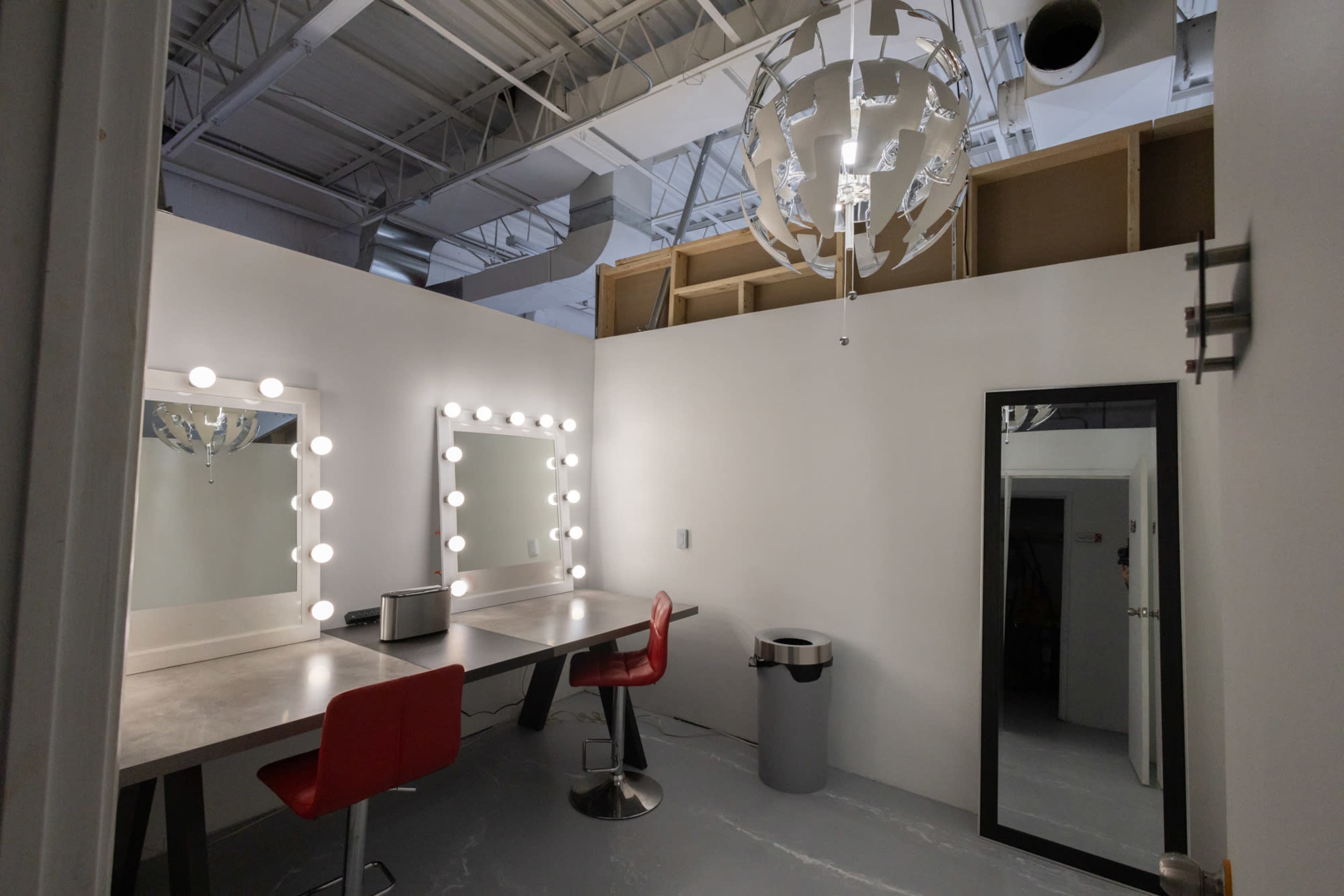 The image shows a modern dressing room with a large mirror framed by light bulbs, a table with red chairs, and a large mirror on an adjacent wall.