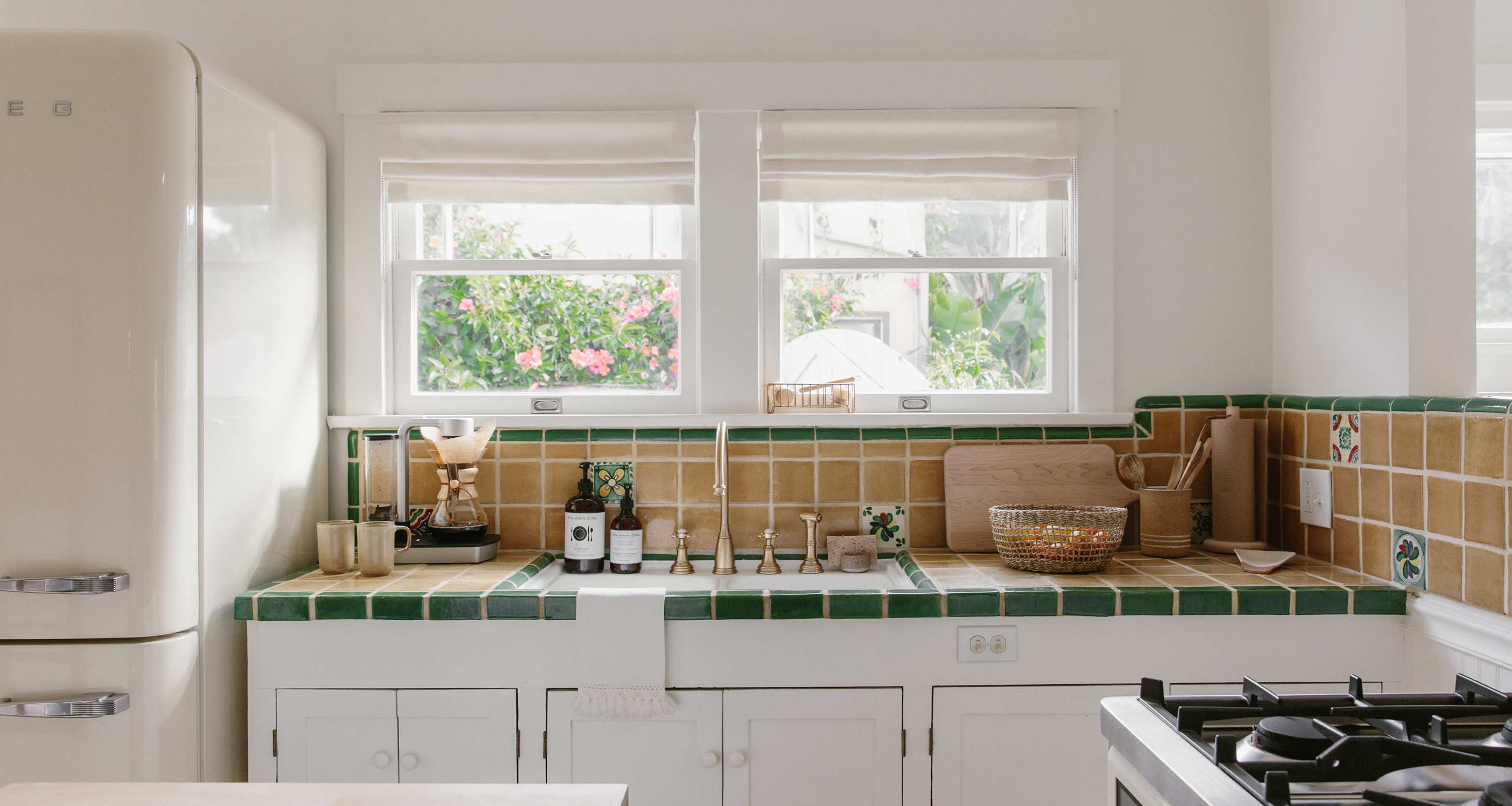 A kitchen with a tiled countertop, a vintage refrigerator, and windows revealing a view of greenery outside.