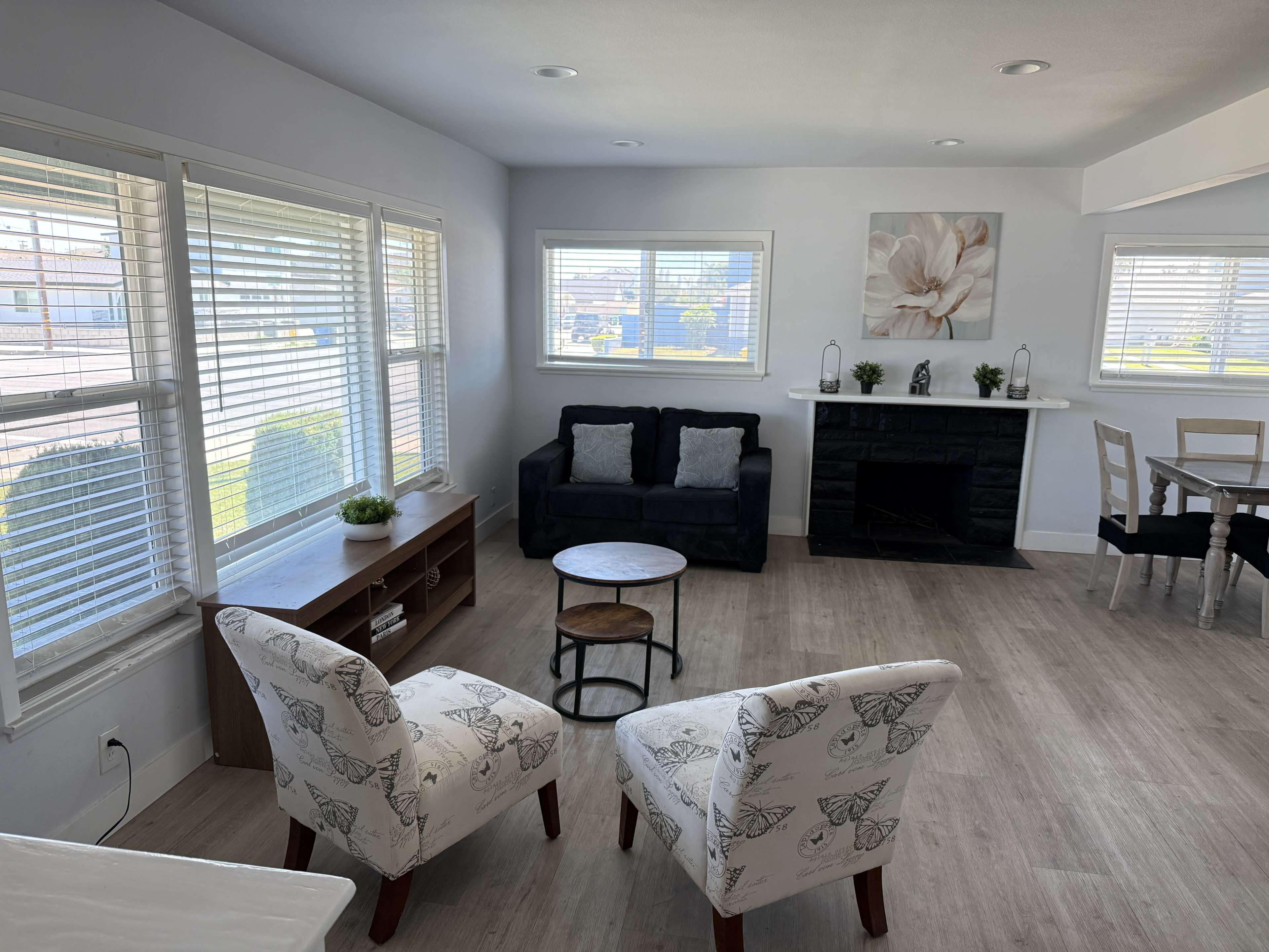 A bright living room features a black couch, two patterned chairs, a round coffee table, and a fireplace, with large windows providing ample natural light.