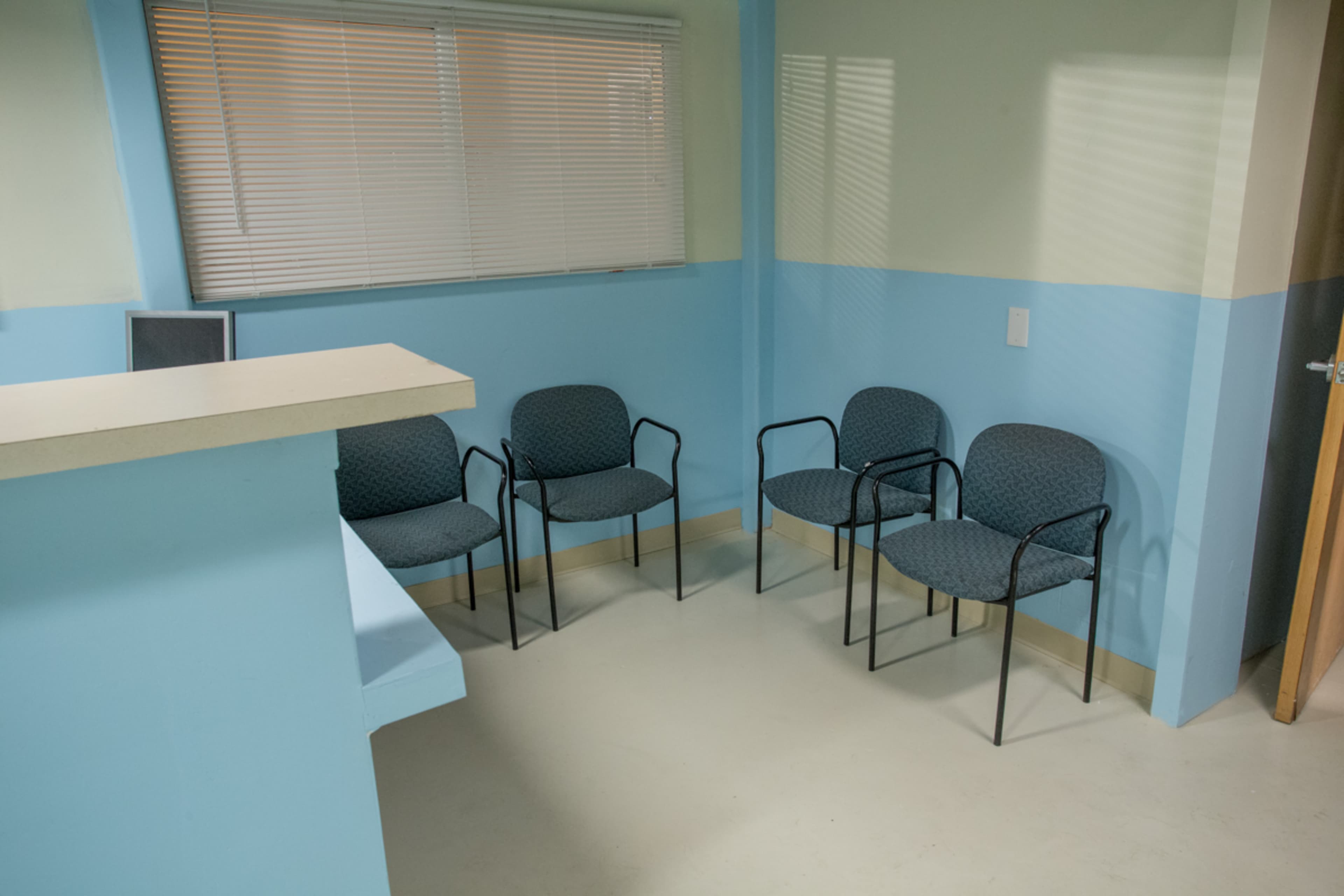 A waiting area contains four chairs and a service counter, with light blue walls and a window with blinds.