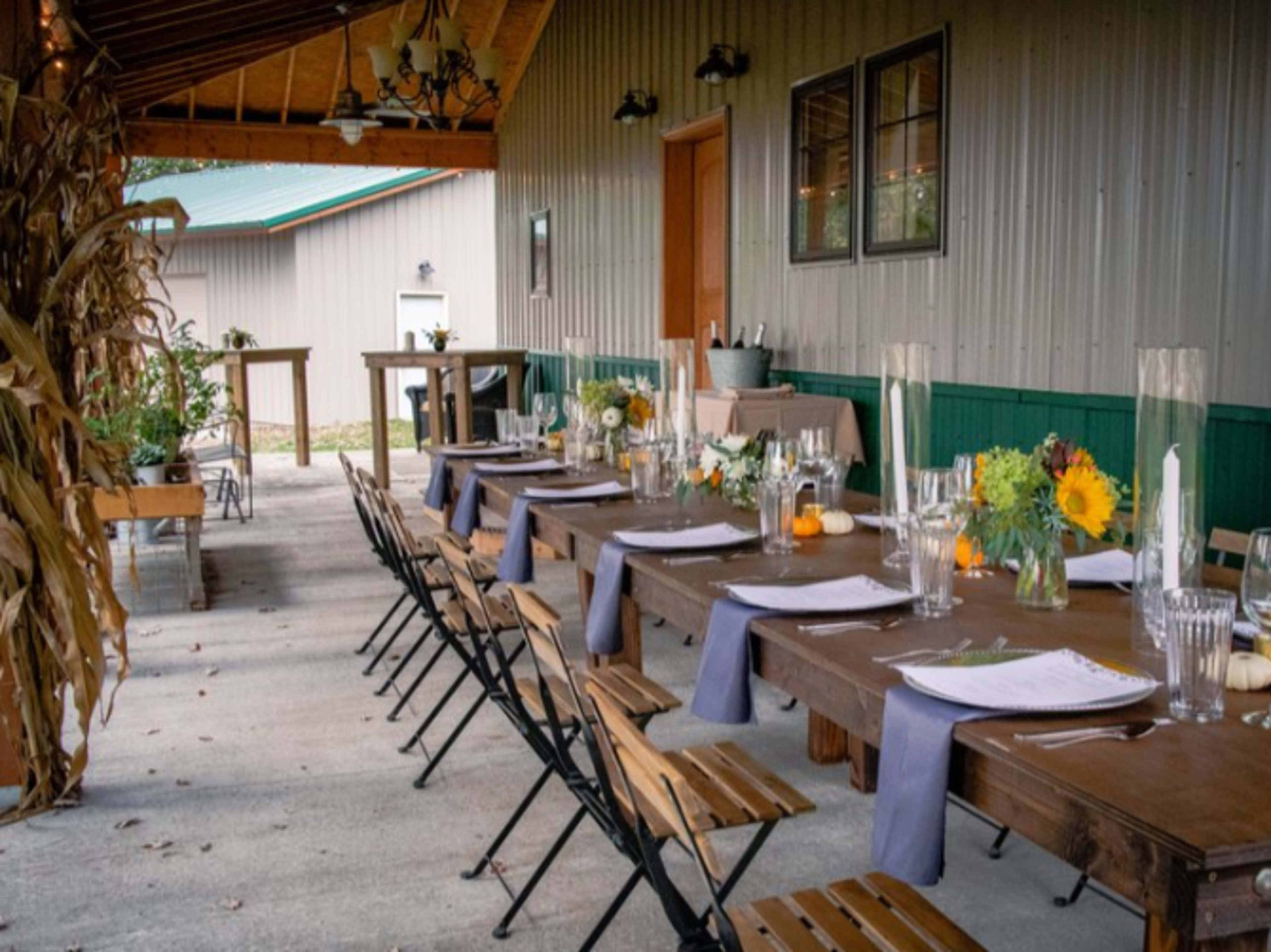 A long table set for a meal is placed outside alongside a building, adorned with flower arrangements and tableware.