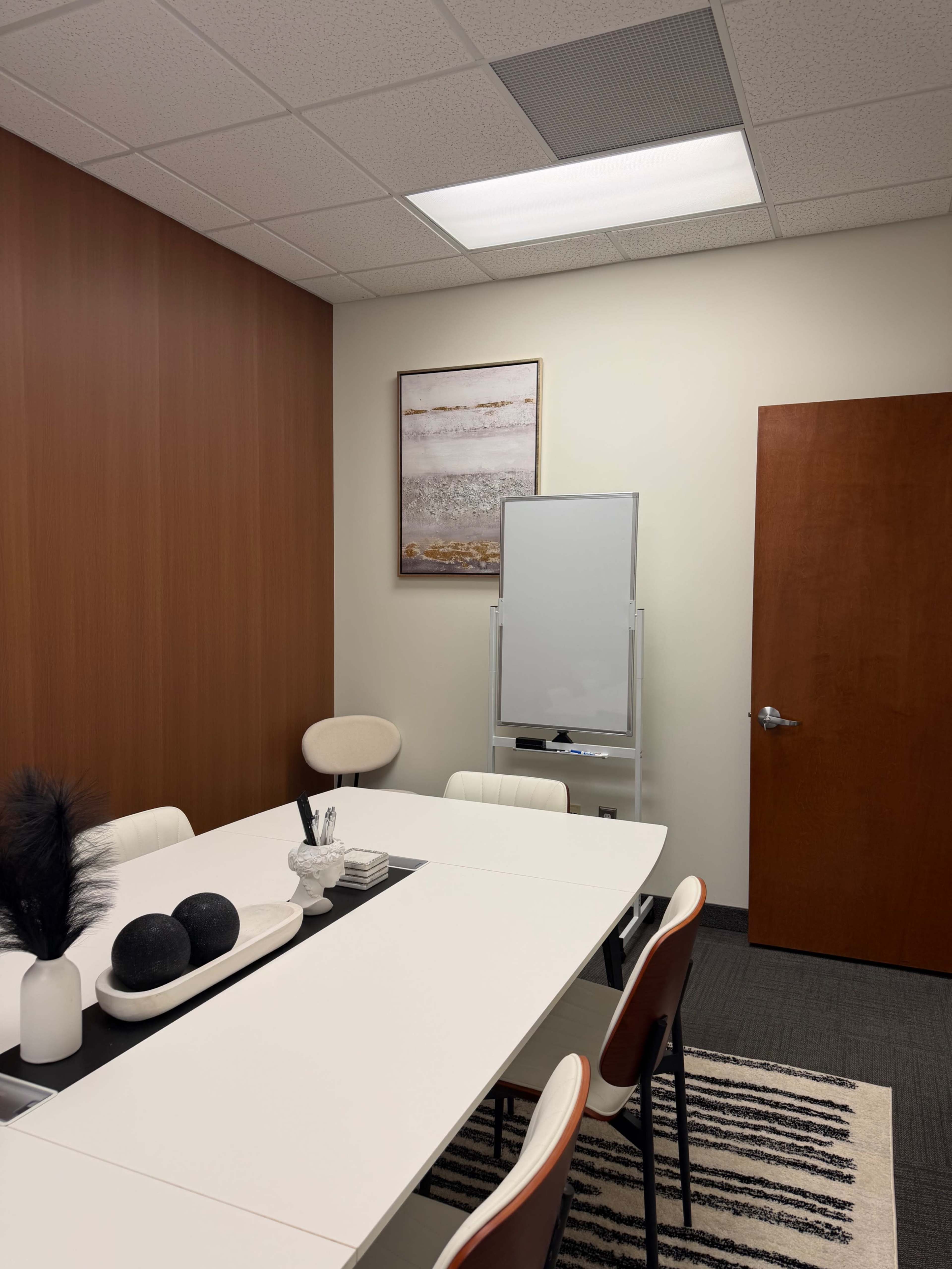 The image shows a modern conference room with a white table, several chairs, a whiteboard, and a piece of art on the wall.