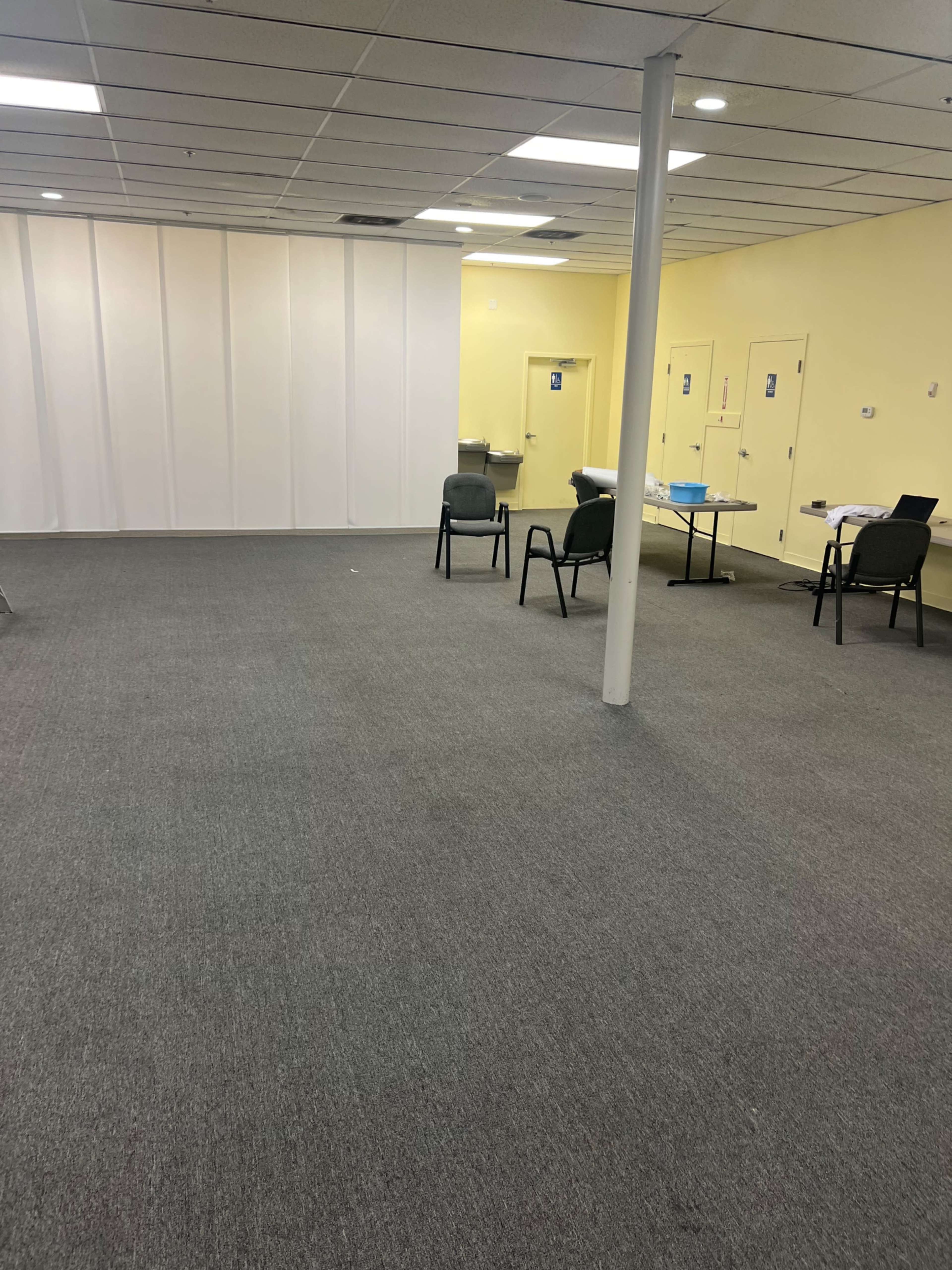 The image shows an empty, sparsely furnished room with yellow walls, a gray carpet, a few chairs, and a table on one side.