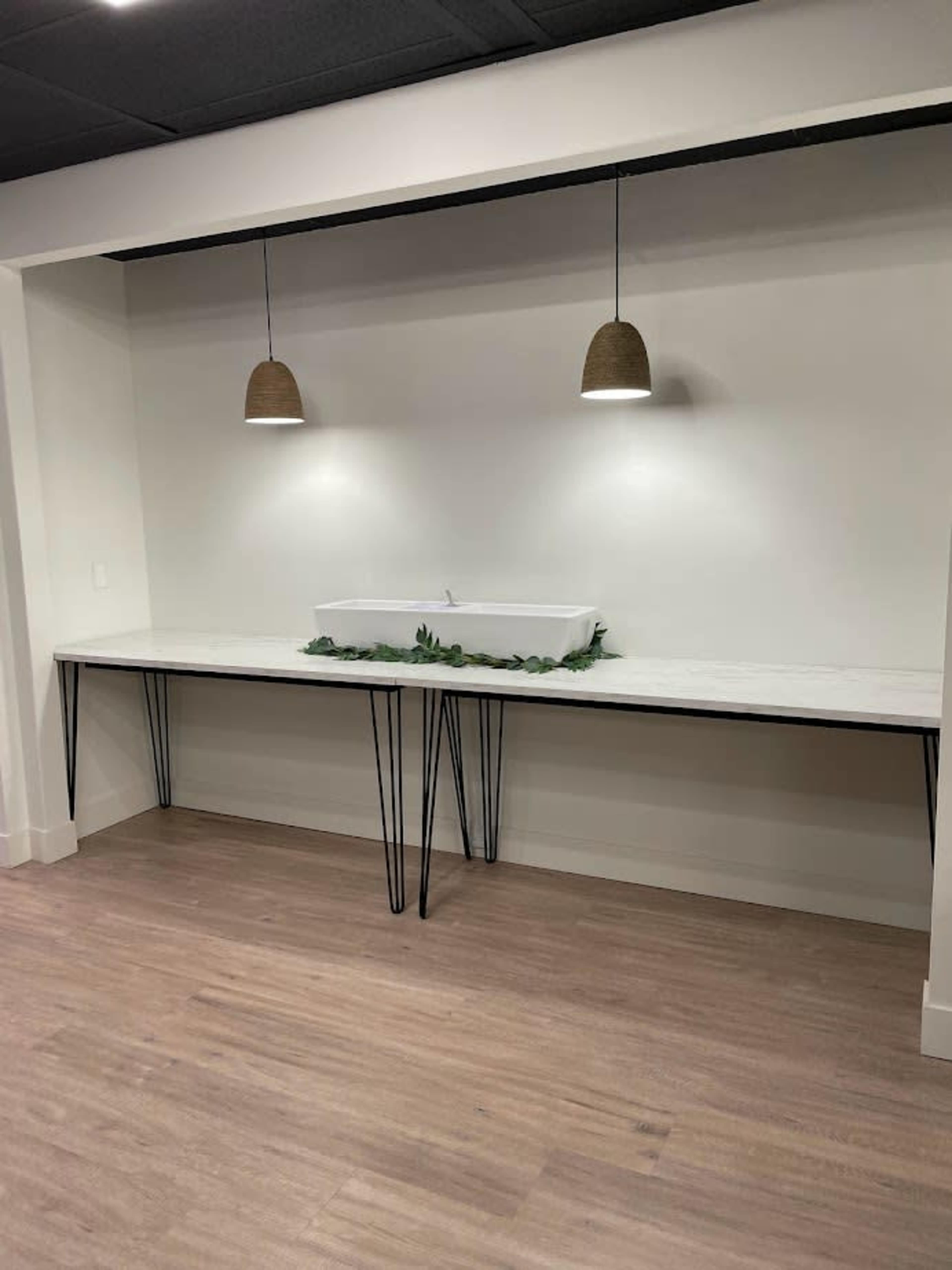 A long, minimalist countertop with a white basin is illuminated by two pendant lights in a spacious room with wooden flooring.