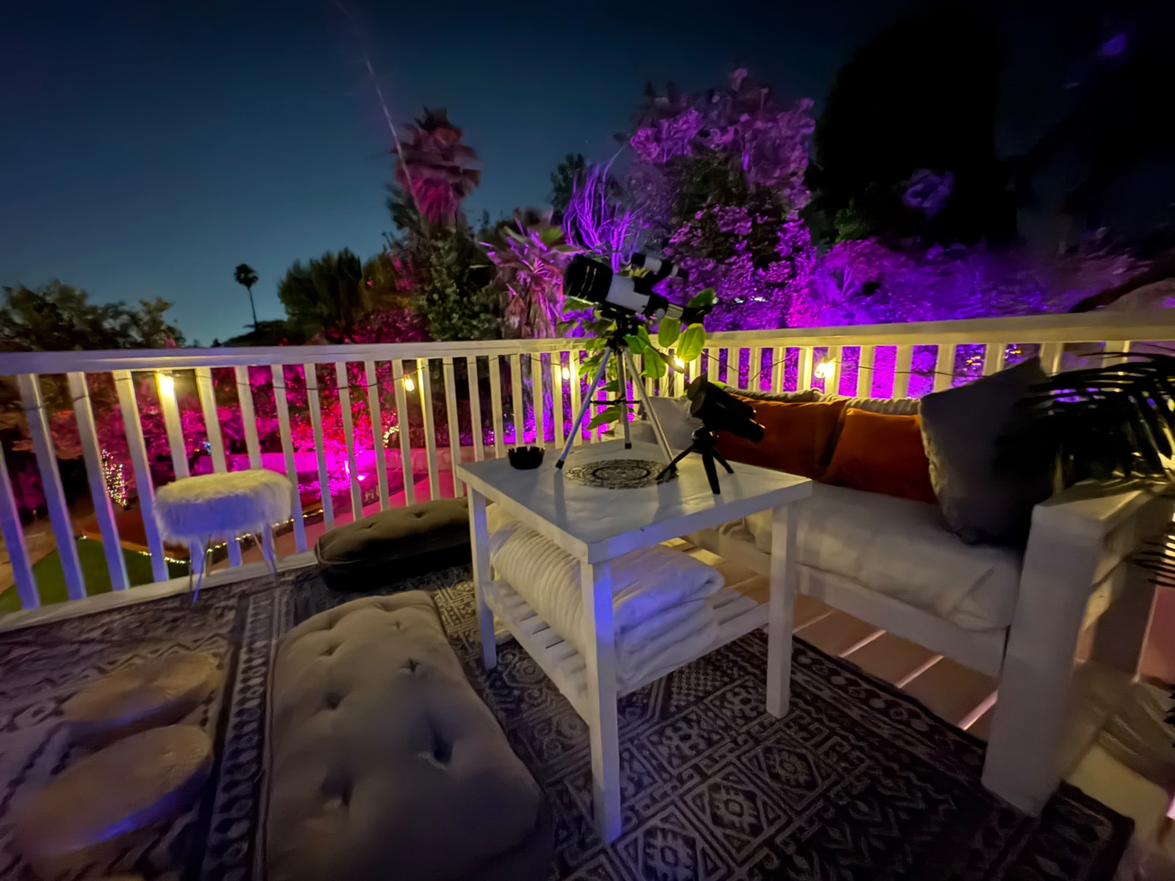 A balcony at night features a white sofa with pillows, a low table, and decorative plants, surrounded by colorful purple lighting and a serene garden view.