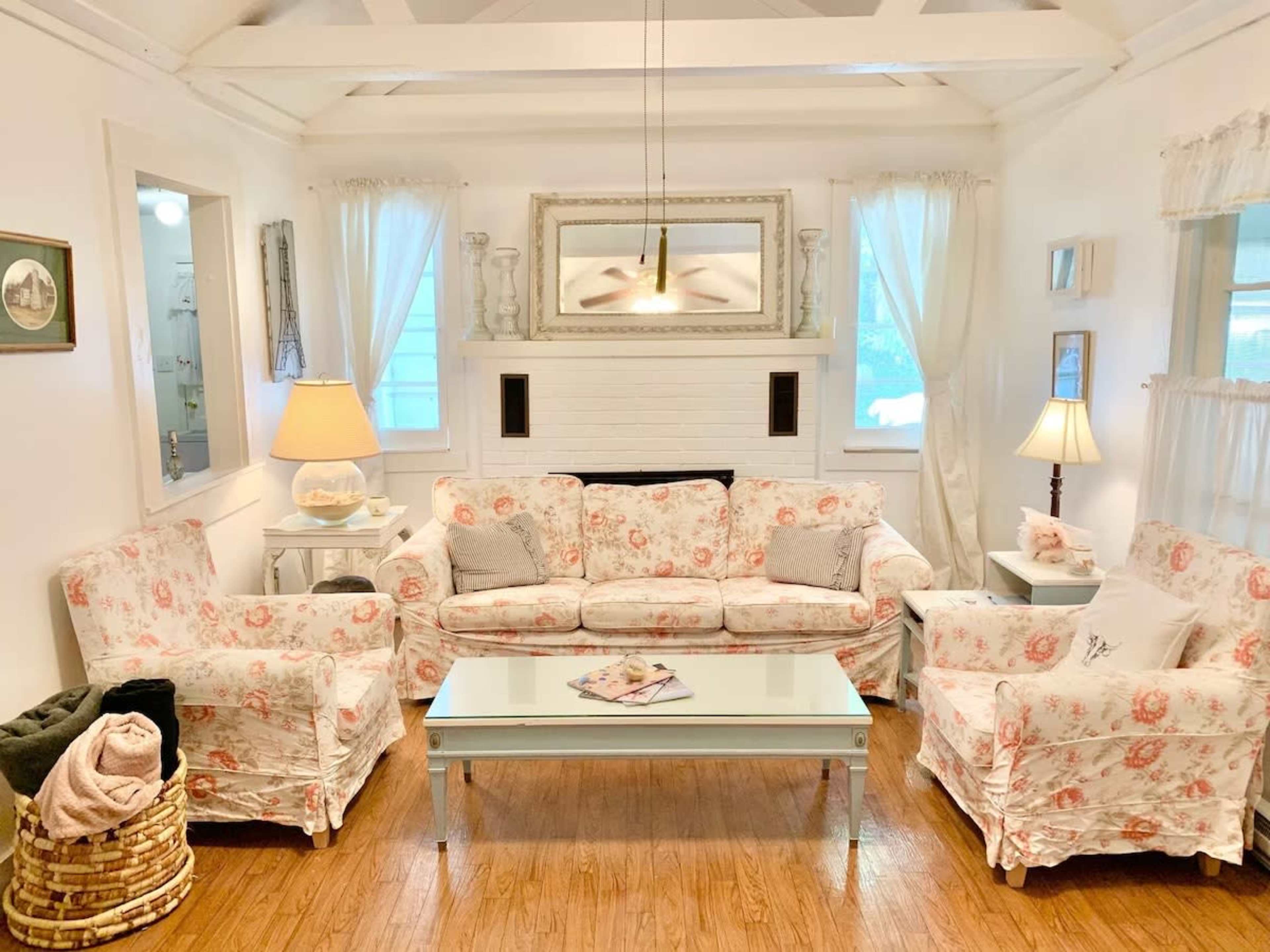A cozy living room with floral-patterned furniture, a glass coffee table, and large windows allowing natural light.