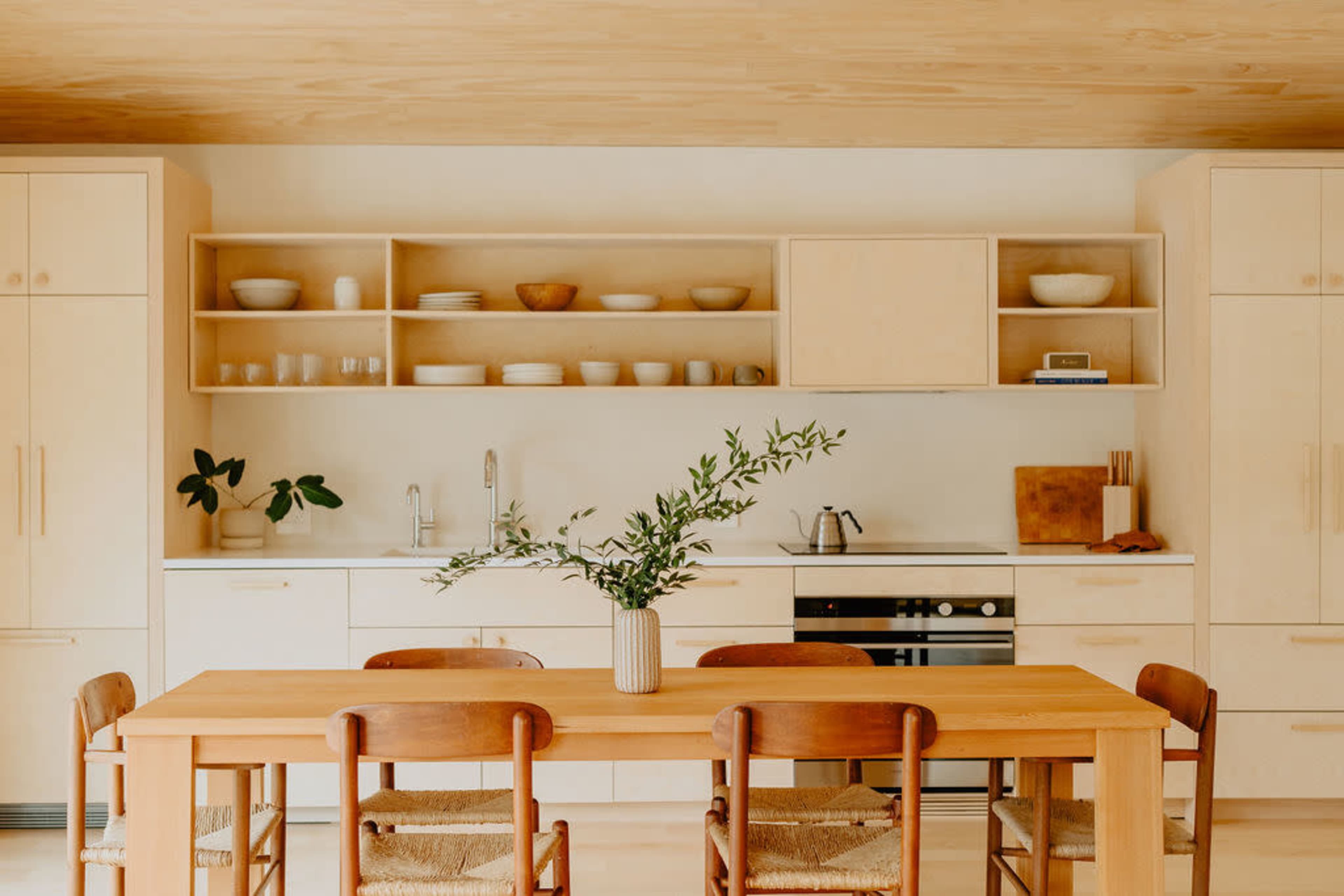 A modern kitchen features light wood cabinetry, a dining table with wooden chairs, and minimalistic decor.