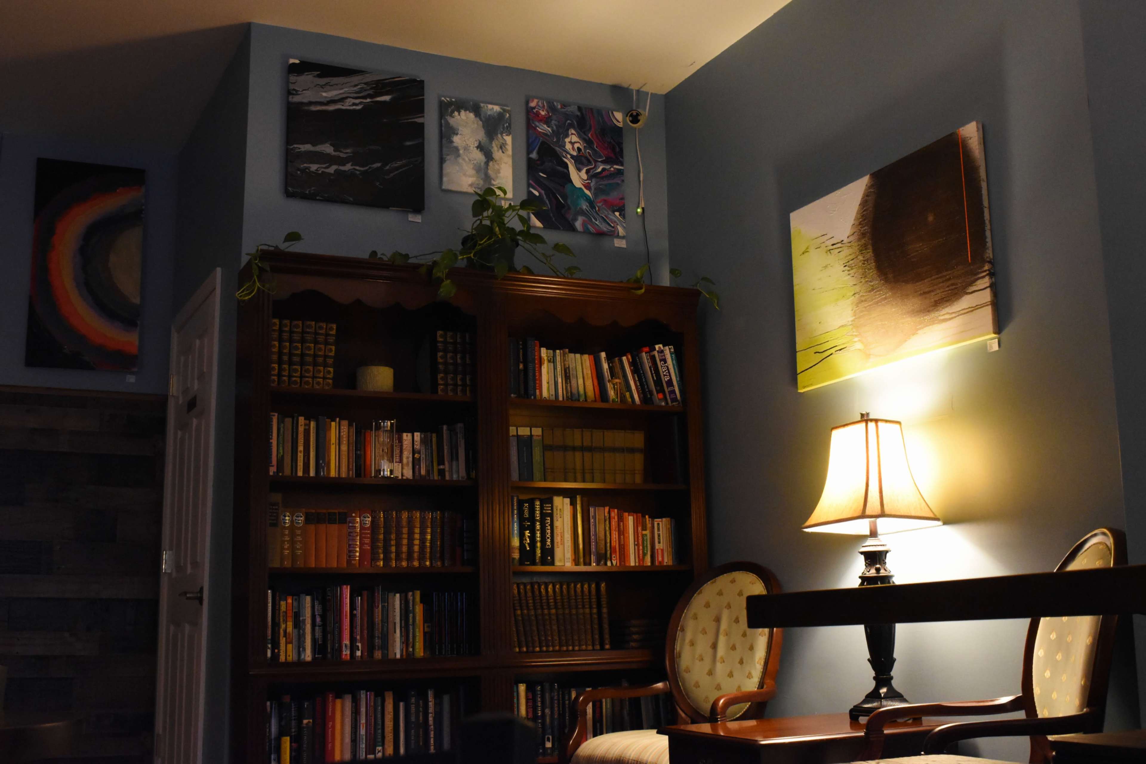A cozy reading corner with a wooden bookshelf filled with books, a lamp illuminating a small table, and wall art displayed above.