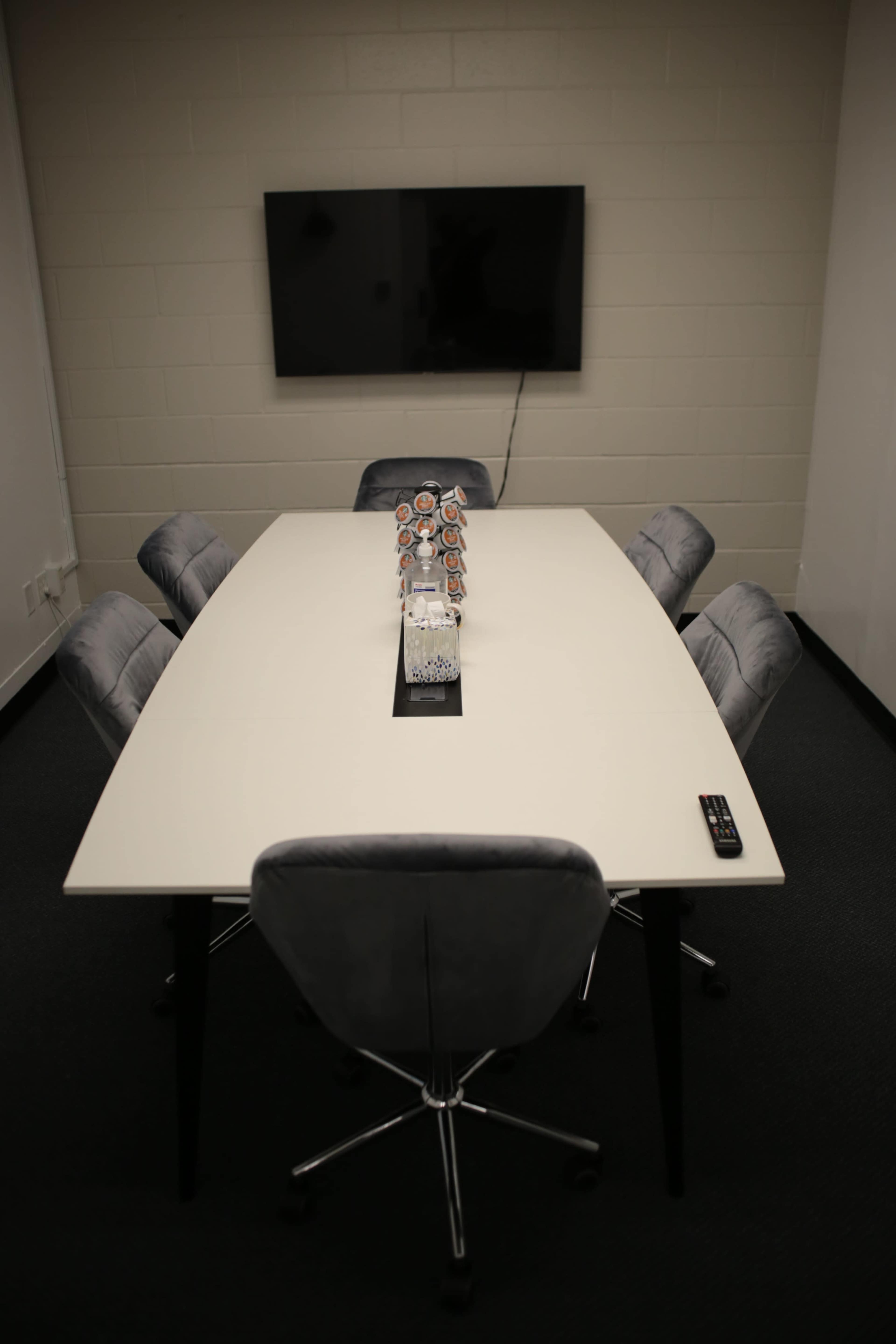 A rectangular conference table surrounded by six cushioned chairs sits in a minimally furnished room with a television mounted on the wall.