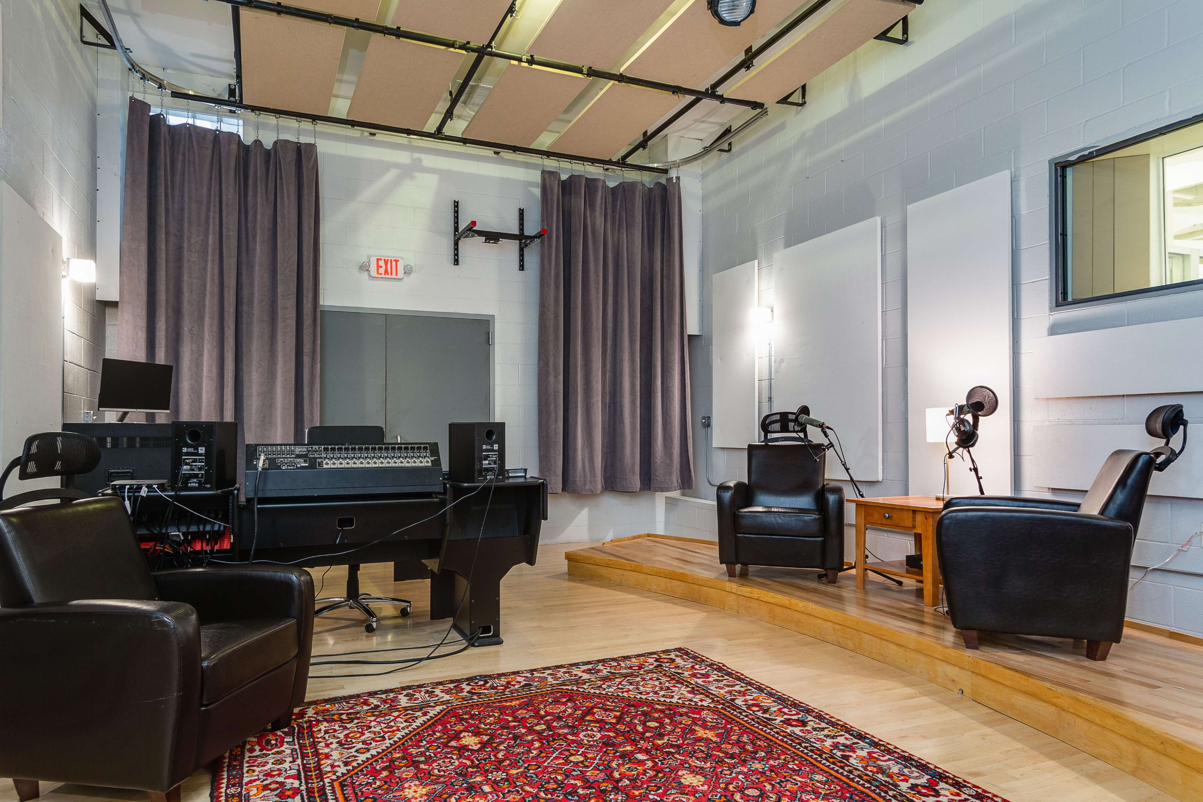 A recording studio with two black leather chairs facing a wooden table and a mixing console, surrounded by soundproof curtains and acoustic panels.