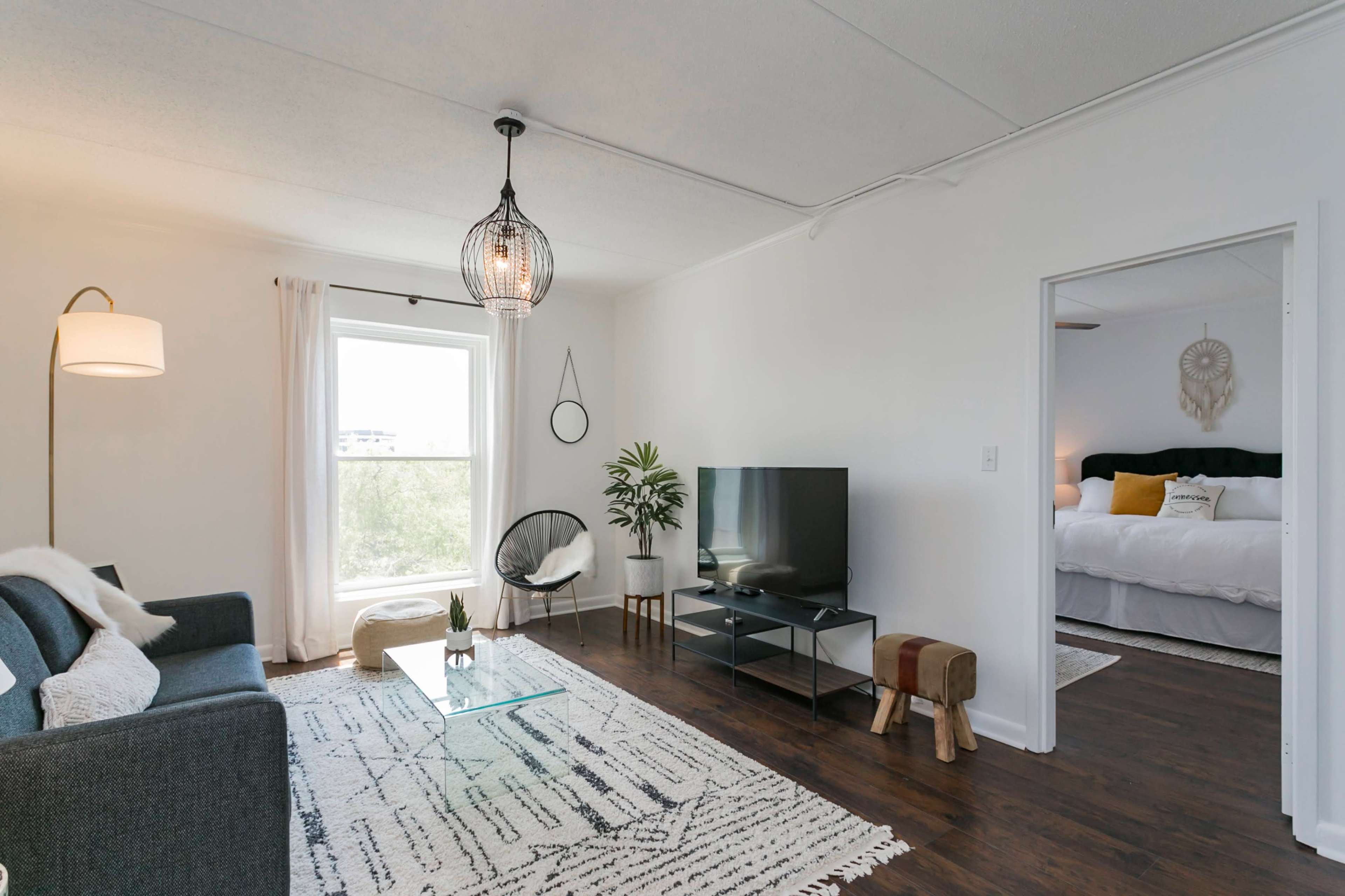 The image shows a bright living room with a grey sofa, a glass coffee table, a television stand, and a doorway leading into a bedroom.