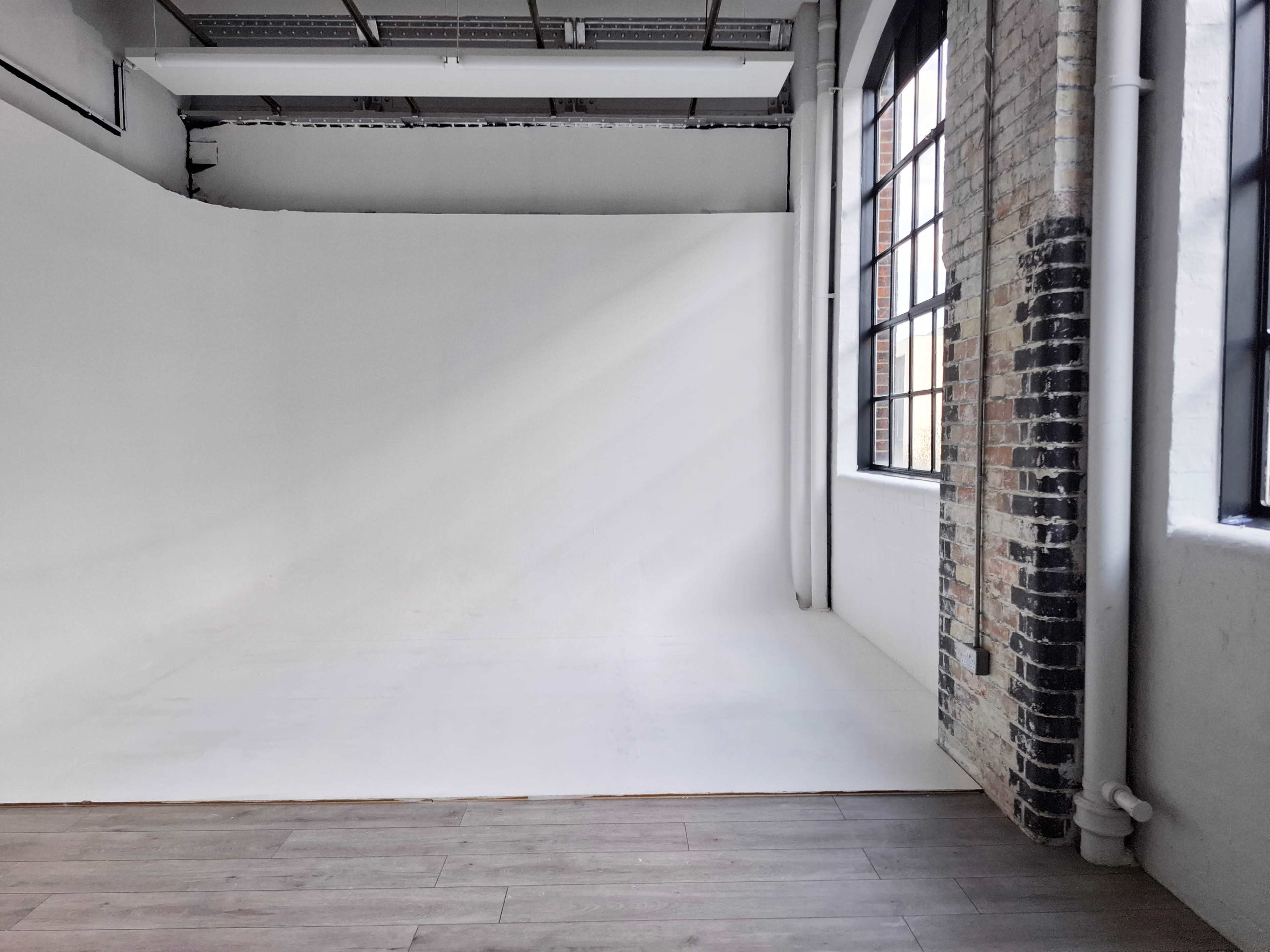 The image shows a spacious studio with a white backdrop, large windows, and exposed brick walls.