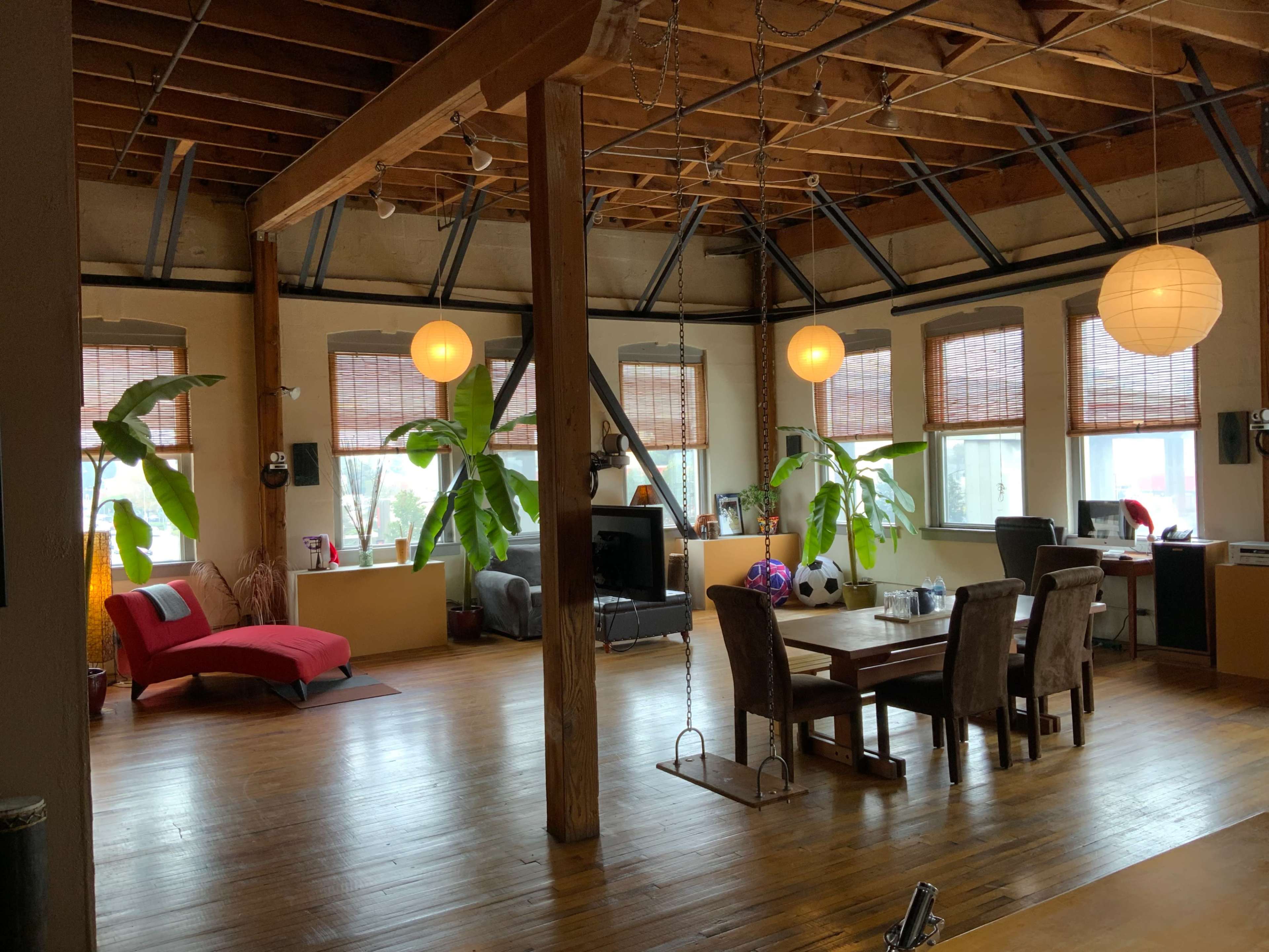 A spacious living area with wooden floors, large windows covered by bamboo shades, and various seating arrangements, including a red couch and a dining table.