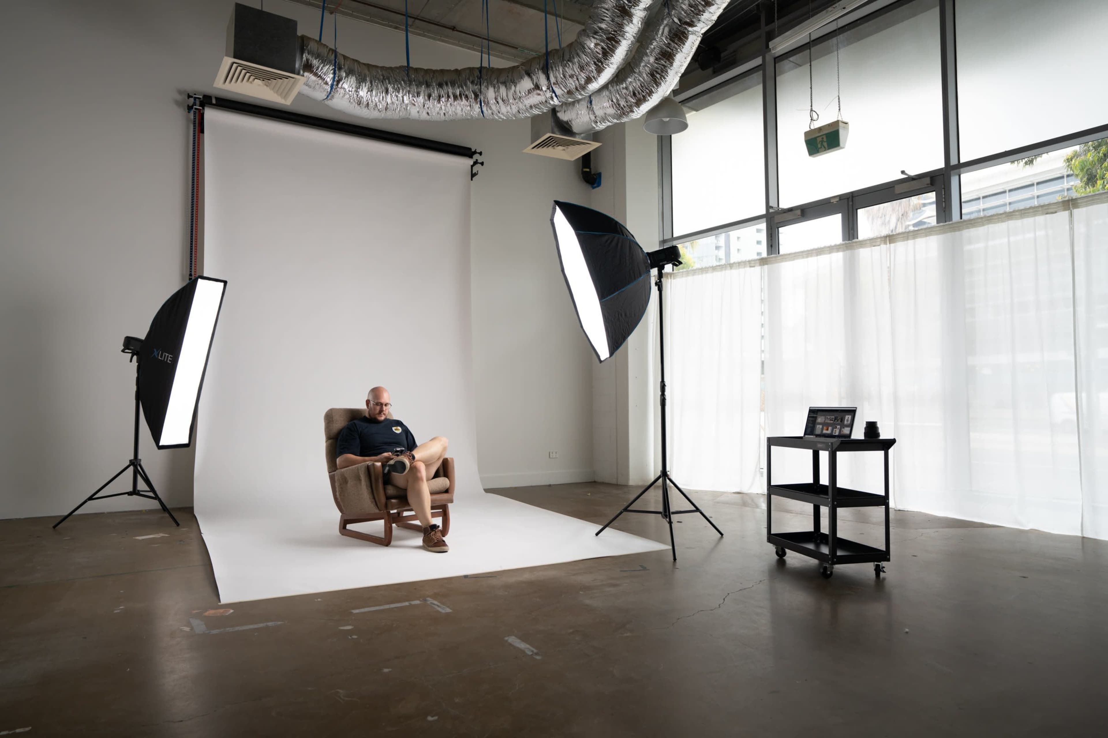 A person sits in a chair on a plain backdrop in a well-lit studio, surrounded by softbox lights and a mobile cart with equipment.