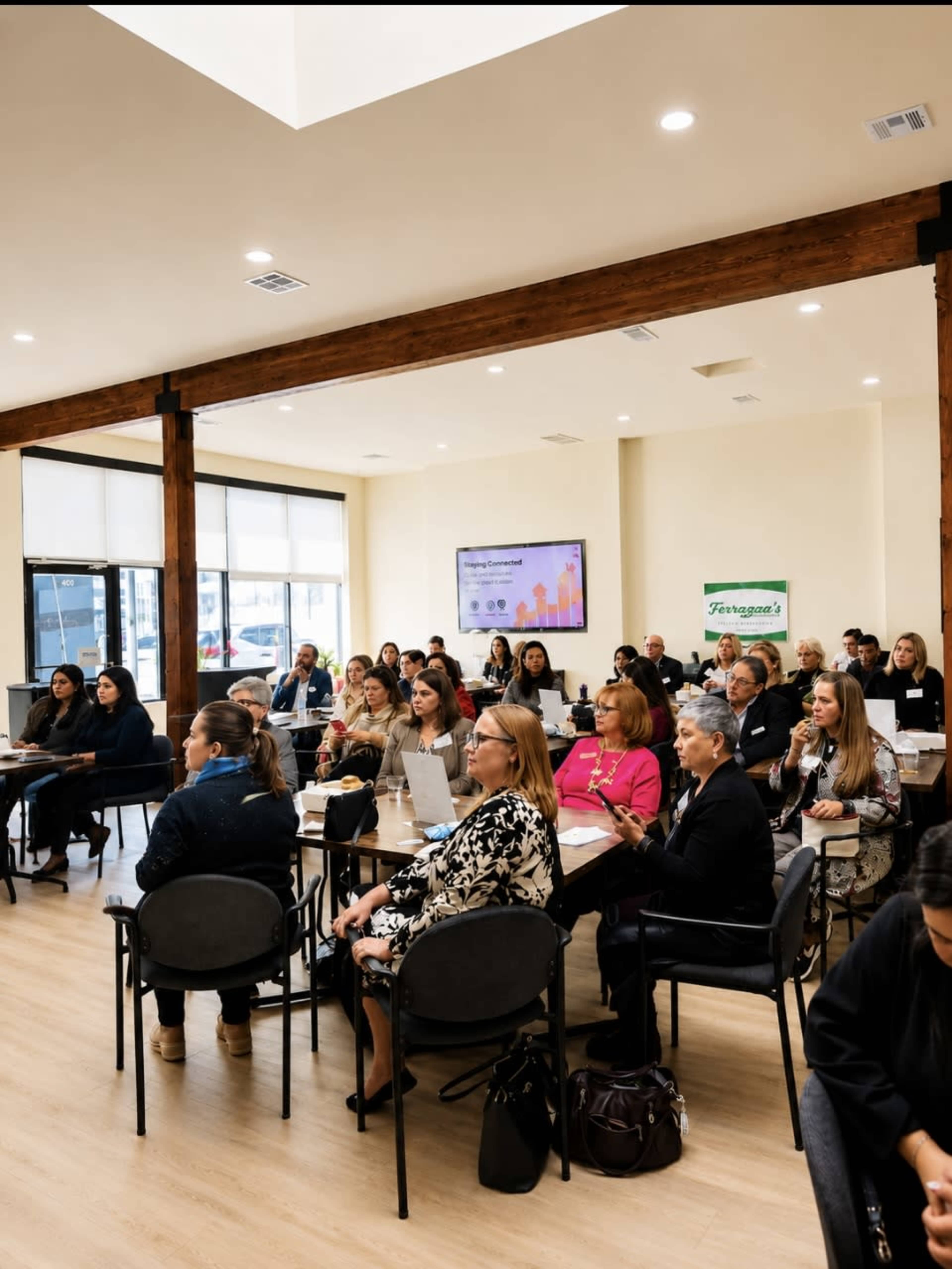 A group of people sit in an indoor meeting space with tables and a presentation screen, engaged in a professional gathering.